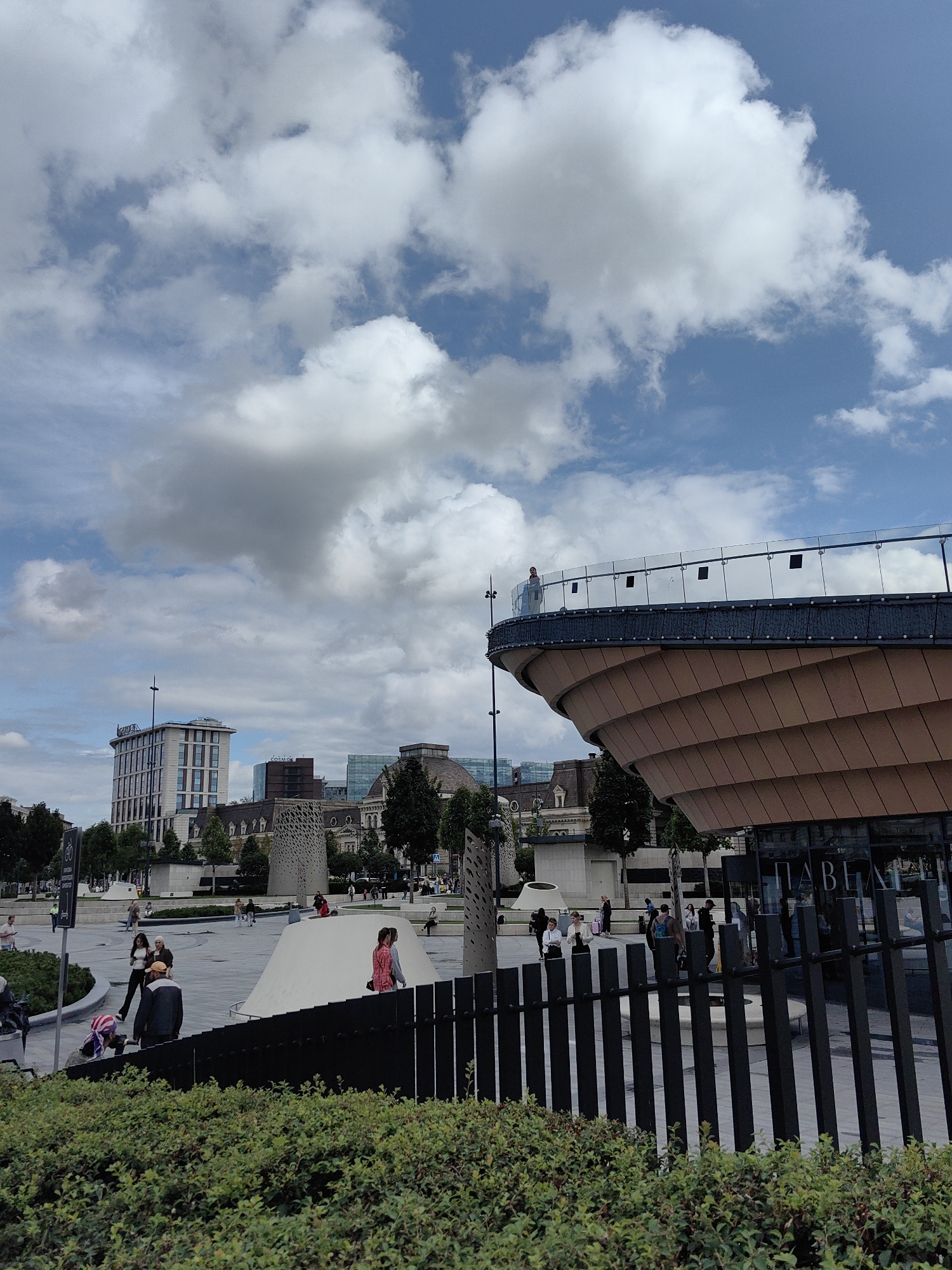 A wide shot captures an outdoor urban plaza on a partly cloudy day. In the foreground, a thick hedge of green bushes lines the bottom of the frame. Behind the hedge, a black metal fence runs diagonally from the right to the left. Beyond the fence, the plaza is paved with light-colored stones and features several abstract white structures, one resembling a large, smooth dome. People are scattered throughout the plaza, some walking, others standing or sitting.
To the right of the plaza, a curved, modern building with a layered, tan facade and a glass-paneled upper deck dominates the mid-ground. A person stands on the upper deck, looking out.
In the background, a cluster of buildings forms the city skyline. The most prominent is a tall, rectangular white building with dark windows and a flagpole on top. Other buildings visible include a glass-fronted structure and a building with a domed roof. Trees are interspersed throughout the scene, with more lining the edges of the plaza. The sky above is a bright blue with numerous white cumulus clouds. The overall impression is of a contemporary public space within a city.