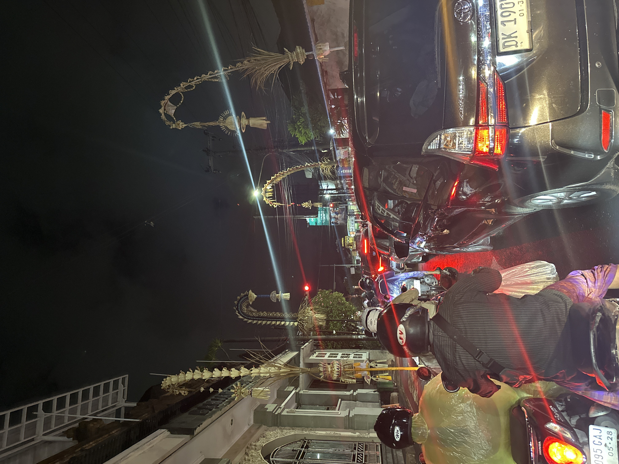 A street at night in North Kuta, Indonesia, is depicted with moderate traffic congestion. Several vehicles, including cars and motorcycles, are stationary or moving slowly. On the left side of the street, a building with traditional Balinese architectural elements is visible, adorned with decorative arches made of palm fronds and flowers. These arches, known as "penjor," are common during Balinese holidays and celebrations. Streetlights and vehicle lights illuminate the scene, creating a dynamic interplay of light and shadow. The sky is dark, indicating nighttime. The road surface appears wet, suggesting recent rain. The overall atmosphere is one of a bustling urban environment during a festive period.