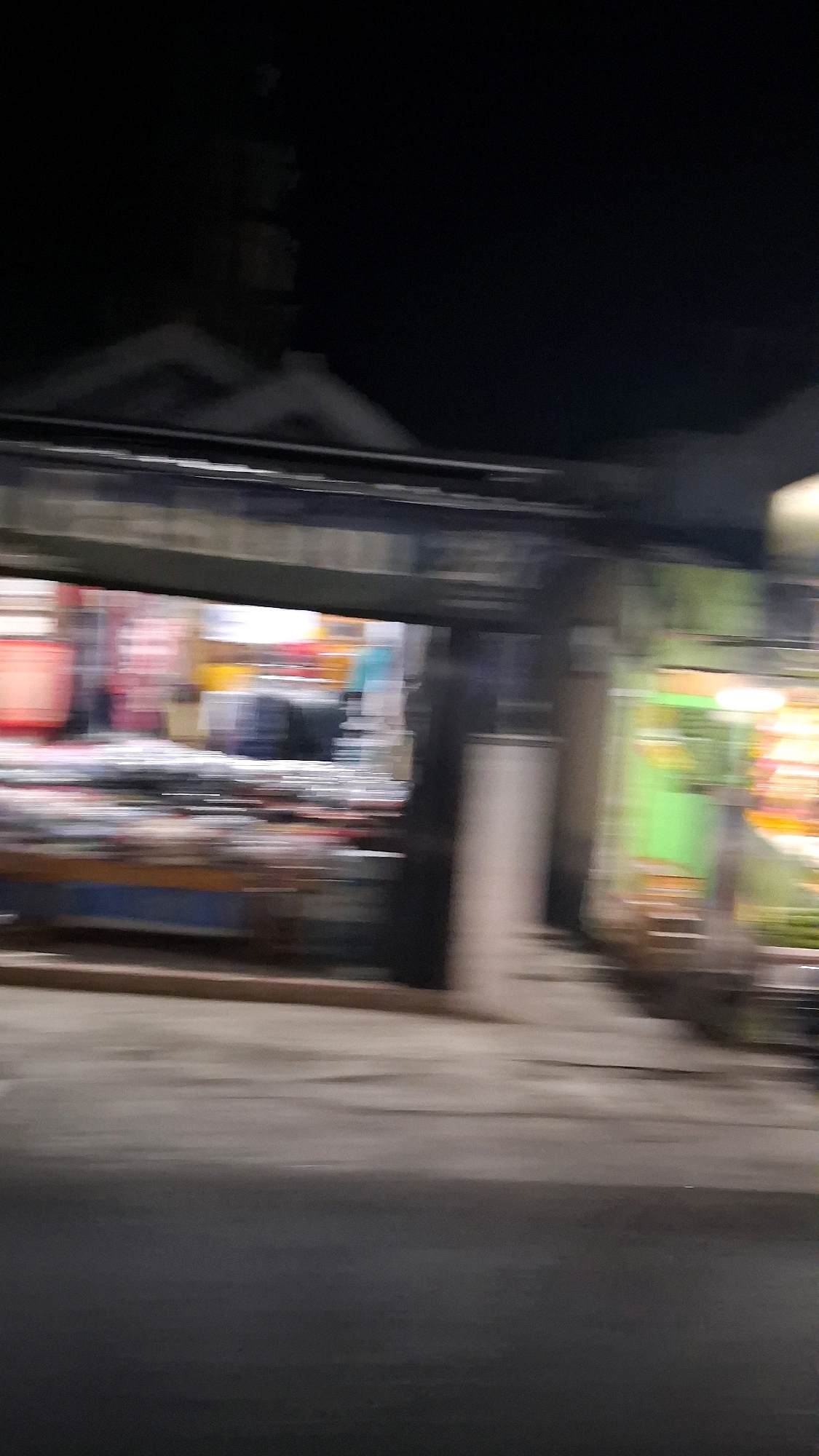 The image depicts a blurry, nighttime scene of a market stall in Berbah, Indonesia. The stall is illuminated by bright lights, casting a colorful glow on the goods displayed. The blurriness suggests that the photo was taken while moving, possibly from a vehicle passing by. The stall appears to be selling various items, with a mix of red, yellow, and blue colors visible. In the background, the roofline of a building is faintly discernible against the dark sky. The overall impression is one of a bustling market scene, captured in a fleeting moment.