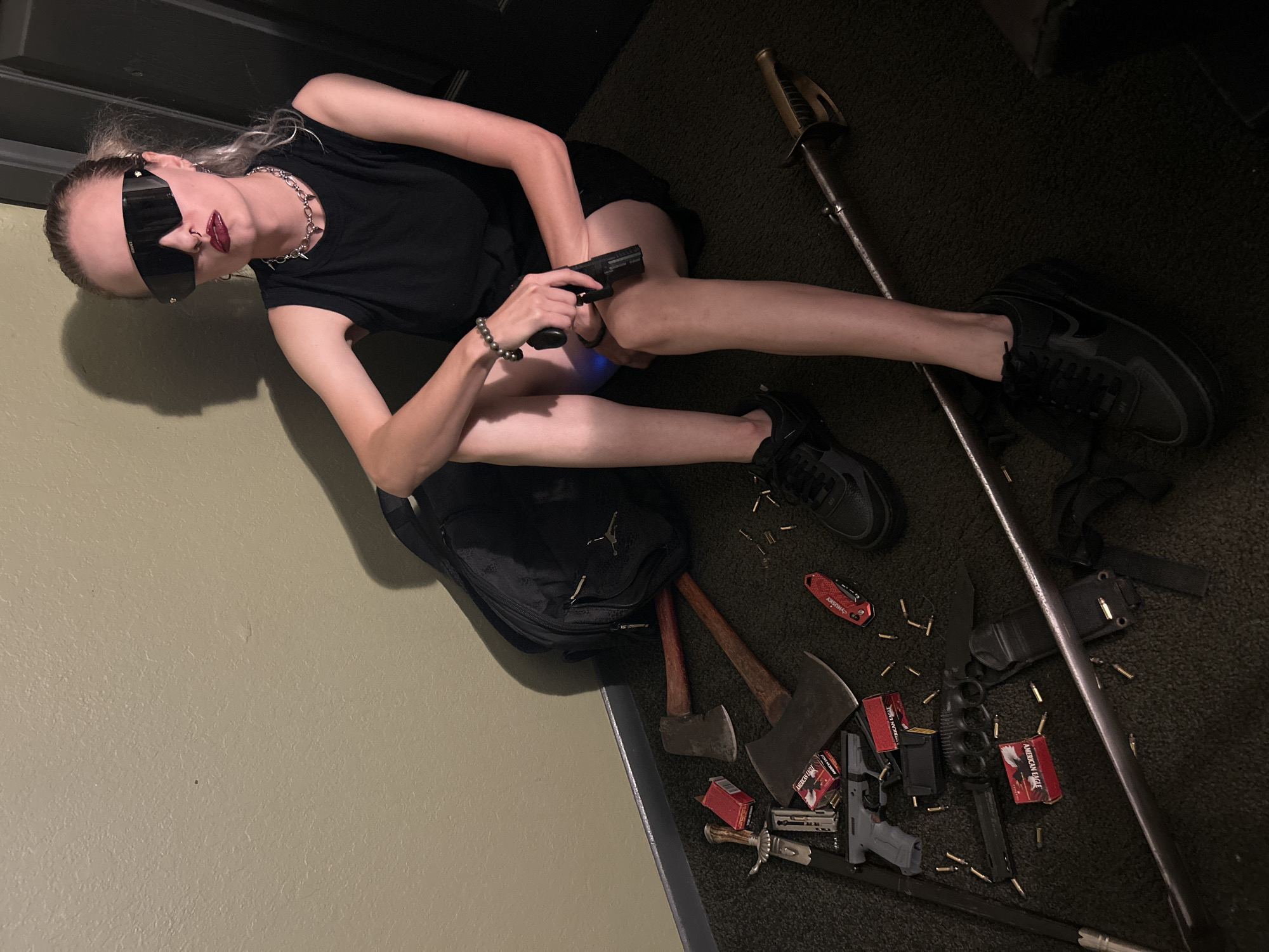 A young individual is captured in an indoor setting, sitting on a dark, possibly carpeted floor with a light tan wall to their left. The subject, likely female, has light-colored hair and is wearing large, dark, rectangular sunglasses that obscure much of their face. They are dressed in a black t-shirt, a black skirt or shorts, and black sneakers. A silver spiked choker necklace and a nose ring are visible, along with dark lipstick. A black bead bracelet adorns their right wrist.
The individual is holding a black semi-automatic handgun in their right hand, with their left hand resting on their knee. Around them, an extensive array of weapons and ammunition is scattered on the floor. These include a black backpack featuring a Jumpman logo, several axes or hatchets with wooden handles, and multiple boxes of "AMERICAN EAGLE" brand ammunition. Numerous loose ammunition casings and bullets are spread across the dark carpet.
Additional visible armaments include at least two other handguns (one gray), several knives of various types such as a butterfly knife, a large fixed-blade knife in a sheath, and a smaller ornate dagger. A red folding utility knife, branded "HUSKY," is also present. Long weapons consist of a decorative sword or saber with a brass hilt and a dark-colored staff or pole. A subtle blue light emanates from beneath the subject's right leg. The scene appears to be a posed photograph showcasing this collection of items, located in an unspecified interior space within Cape Coral, United States of America.