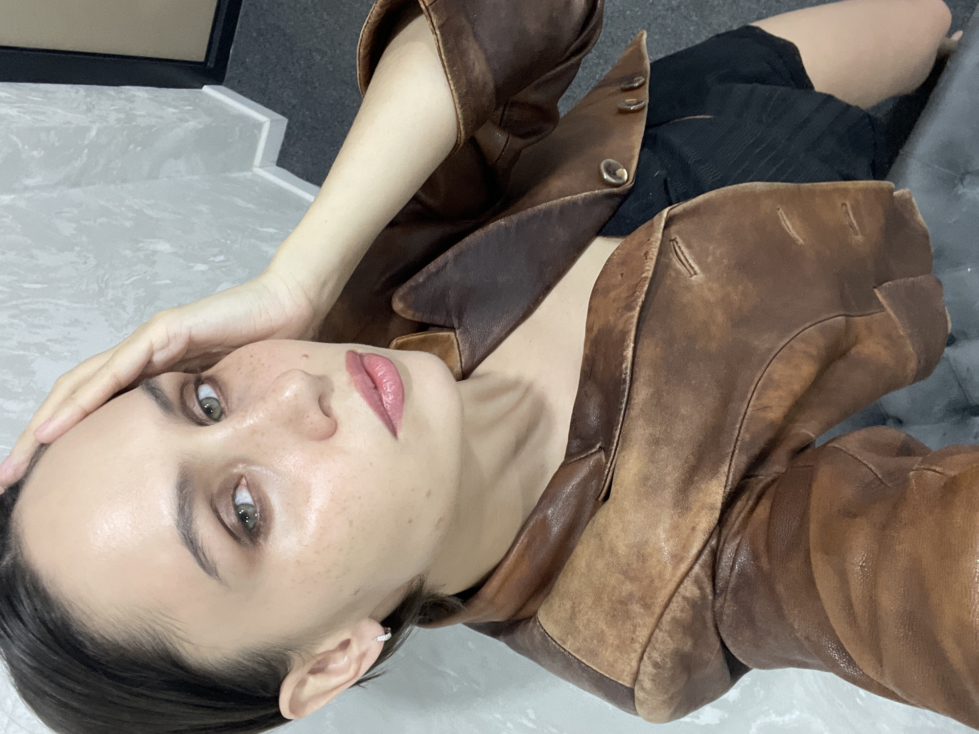 A woman with dark hair and light skin is lying on a light-colored surface. She is wearing a brown leather jacket and a black garment underneath. The background shows a light-colored wall or floor with a hint of a dark object. The scene does not provide enough information to identify a specific location, let alone one in Izhevsk, Russia.