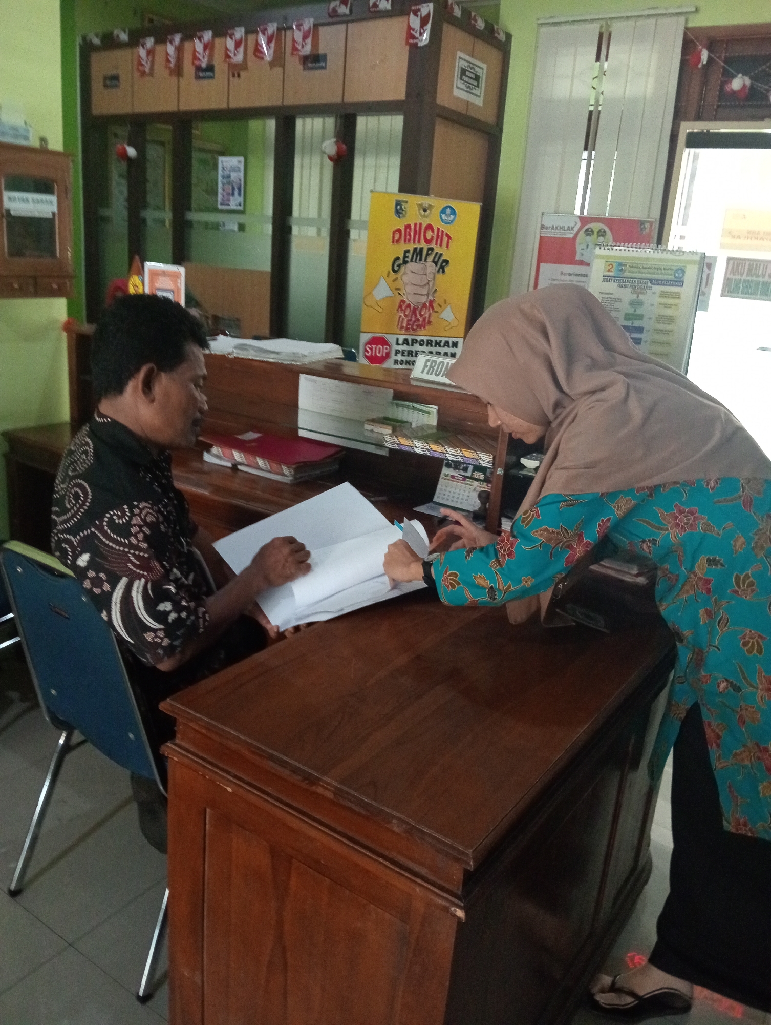 The image depicts an indoor scene, likely an office or administrative area, possibly in Demak, Indonesia, based on the Indonesian language text visible on some of the objects. Two individuals are present: a man in a black batik shirt and a woman wearing a hijab and a blue and red floral patterned top. They are both interacting with documents on a large wooden desk. The man is seated and appears to be reviewing a document, while the woman is standing and leaning over the desk, also engaged with the papers.
The environment is characterized by wooden furniture, including the desk and cabinets lining the wall. A yellow poster with Indonesian text and graphics, including a hand icon and the word "STOP," is visible on the left. Other documents, folders, and what appears to be a calendar are also on the desk and shelves. The lighting suggests an indoor setting, possibly with natural light coming from a window partially obscured by blinds in the background. The overall impression is one of an active workspace where administrative tasks are being carried out.