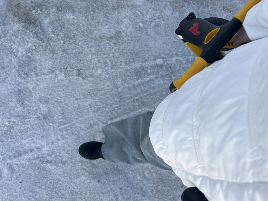 A first-person perspective image captures an individual standing or walking on a textured, icy, and snowy surface in Chaikovsky, Russia. The subject is partially visible, wearing light blue/grey jeans and a black boot on the left foot. A white puffer jacket covers the lower torso and thighs. The individual's right hand grasps a yellow and black object, consistent with the handlebars and frame of a child's balance bike or similar ride-on toy, which features a red squirrel logo on its black component. The ground consists of unevenly distributed ice and packed snow.