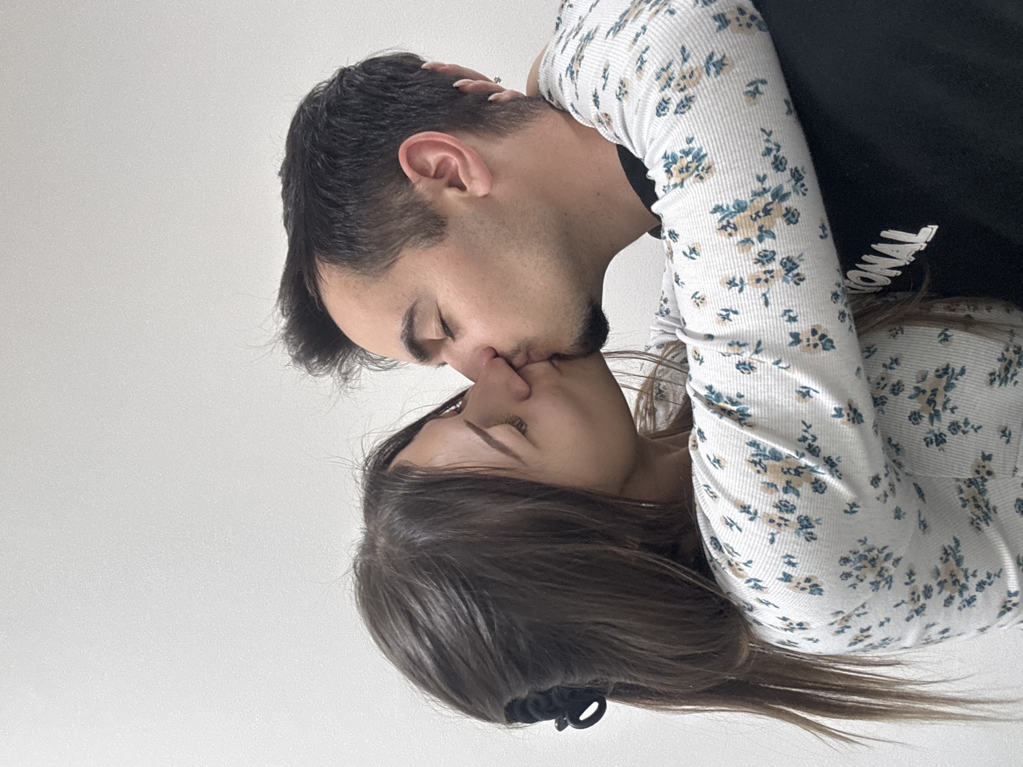 A man and a woman are kissing. The man has dark hair and is wearing a black shirt and a white floral-patterned shirt. The woman has long brown hair and is wearing a floral-patterned shirt that matches the man's. The background is a plain white wall. The image does not contain information to determine its location in Sofia, Bulgaria.