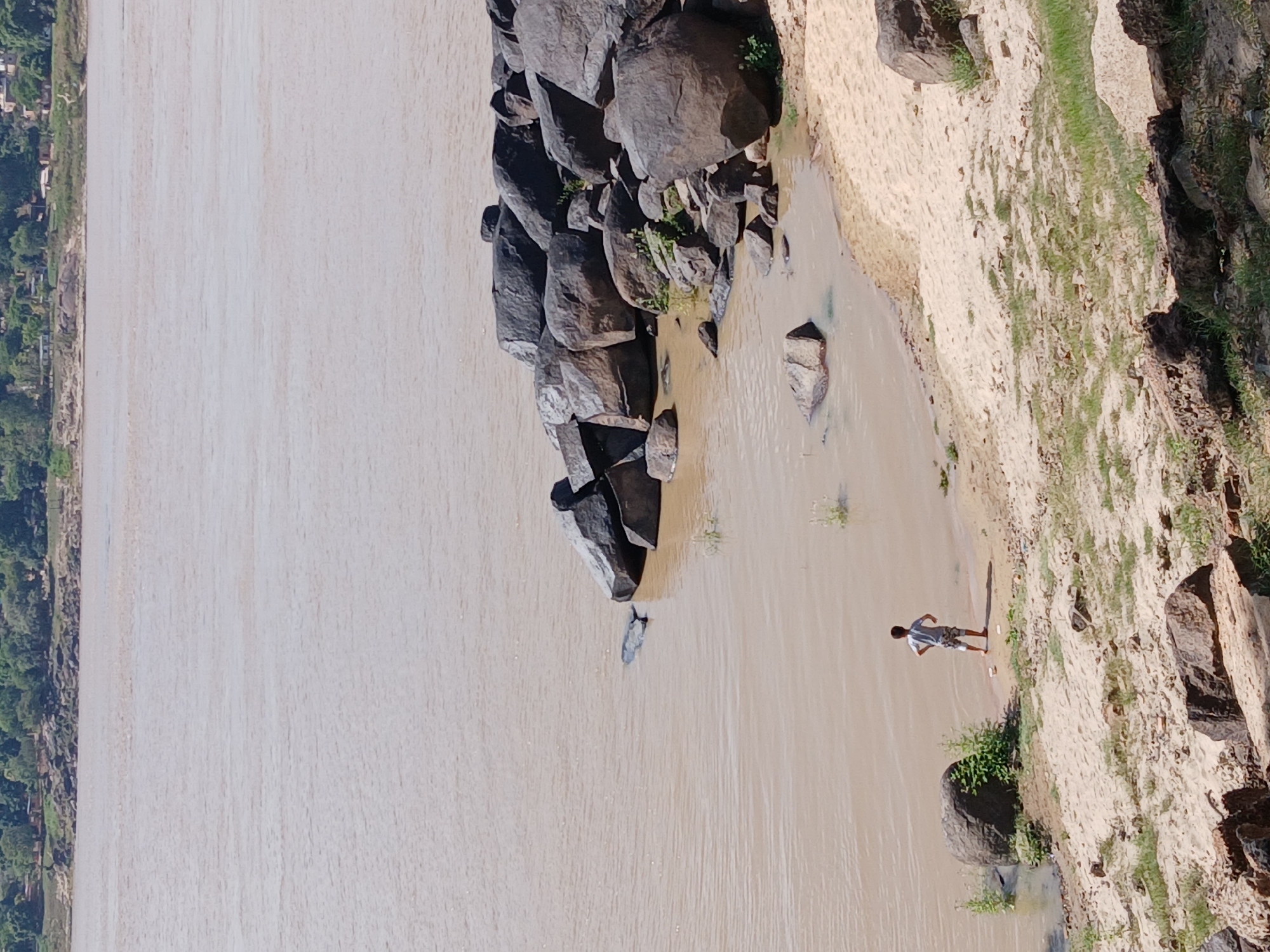 The image shows a wide, muddy river bordering a sandy bank with rocks and vegetation. A person stands on the bank near the water's edge, casting a long shadow. The opposite bank is visible with dense trees. The river is the Orinoco River in Ciudad Bolivar, Venezuela.