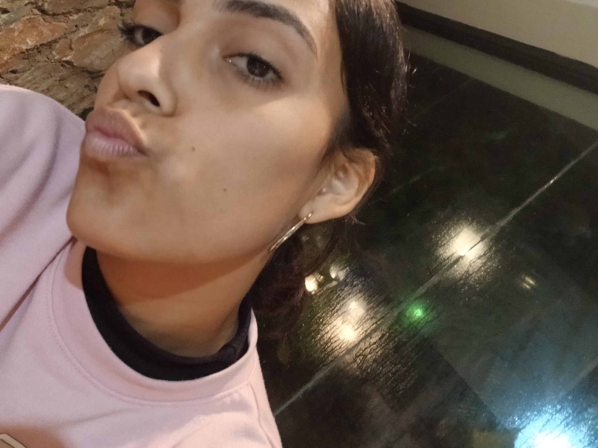 A young woman with dark hair and light skin is taking a selfie. She is wearing a pink sweatshirt with a black collar. Her lips are pursed, and she is looking slightly upwards. In the background, there is a window with reflections of lights and what appears to be rain on the glass. To the left of the woman's face, there is a textured surface that resembles tree bark. The scene is likely indoors, possibly at a restaurant or bar with a view of an outdoor area at night. There are no other people or significant objects visible in the frame. The specific location of Playa El Agua, Venezuela, cannot be determined from the provided image.
