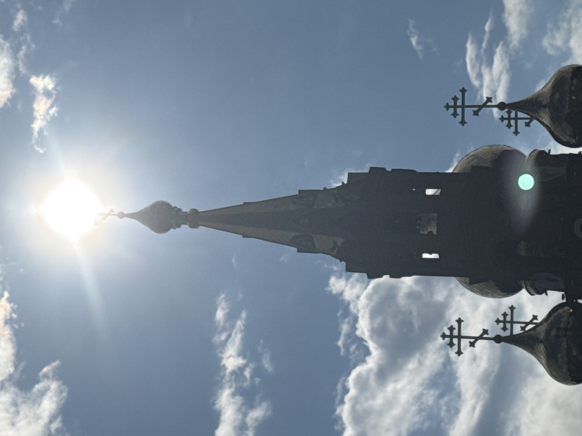 A low-angle, silhouette shot of an Orthodox church spire piercing a bright, sunlit sky in Chaikovsky, Russia. The sun is directly overhead, creating a lens flare with visible rays. Fluffy white clouds drift across a clear blue sky. The dark silhouette of the spire features intricate architectural details, including windows and ornate crosses at the apex and on smaller domes. Several other dark domes with crosses are visible in the foreground and to the sides of the main spire, also silhouetted against the bright sky. One of the domes shows a bright green lens flare reflection. The image captures a moment of spiritual grandeur against a vibrant natural backdrop.