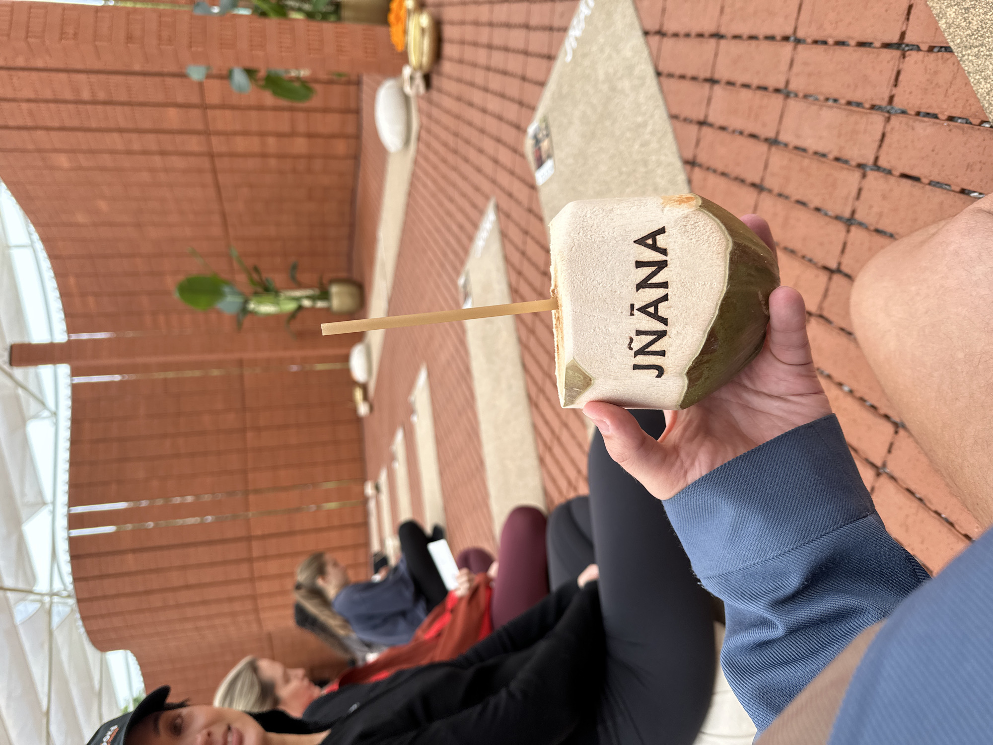 A person's hand holds a halved coconut with a straw inserted, with the word "JÑĀNA" branded on its side, in the foreground. In the background, a group of people are sitting on mats in a yoga class. The location is an outdoor covered area with brick walls and a white canopy ceiling, suggesting an urban setting. The people are dressed in athletic wear, and some have yoga mats and water bottles. The overall atmosphere appears serene and focused. There are plants in pots and some decorative items like small golden bowls and flowers placed on the mats, contributing to a holistic or wellness theme. The flooring is made of interlocking brick tiles. This scene is likely at a wellness center or a retreat in London, United Kingdom.