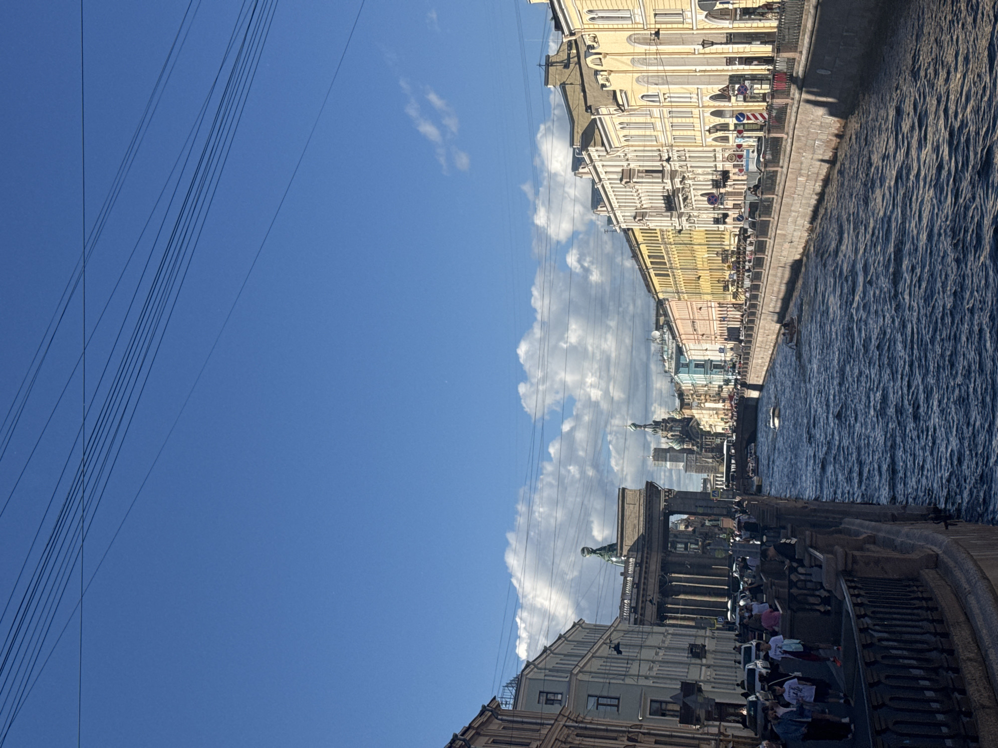 The image captures a view of the Kazan Canal in Saint Petersburg, Russia, on a bright, partly cloudy day. The canal occupies the foreground, with its dark, rippling water reflecting the sky and surrounding architecture. On the left bank, a stone embankment with a black wrought-iron railing lines the canal, with a sidewalk populated by pedestrians. The buildings along this side are multi-story, with varied architectural styles and facades in muted colors, including a pale blue building.
Across the canal, on the right bank, a line of colorful, classic European-style buildings stretches into the distance. These buildings are predominantly in shades of yellow and cream, featuring numerous windows, arched doorways, and decorative architectural elements. The street behind them is visible, with cars parked along the side and more pedestrians walking on the sidewalk, which also features a railing.
In the middle ground, several prominent landmarks of Saint Petersburg are visible. A large, classical colonnade structure, likely part of Kazan Cathedral, is prominent on the left side of the canal. Further down the canal, the distinctive onion domes of the Church of the Savior on Spilled Blood can be seen against the sky, along with other architectural spires. A few small boats are present on the canal, including one motorboat moving away from the viewer. Overhead, a series of dark electrical wires are strung across the bright blue sky, casting faint shadows. The overall atmosphere is that of a bustling urban scene set against a backdrop of significant historical and architectural significance.
