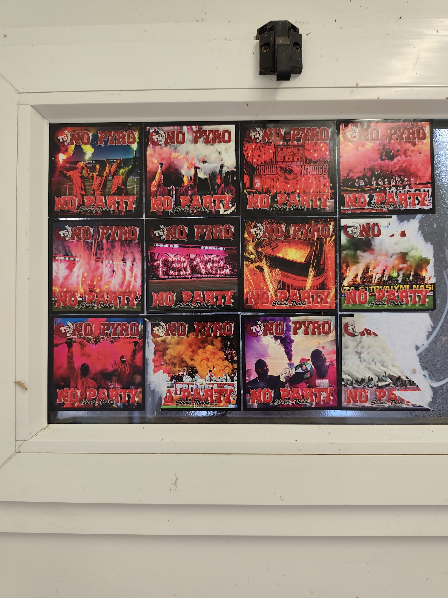 The image shows a display of twelve square stickers, arranged in a 3x4 grid, adhered to a white, painted surface that appears to be a window frame. Each sticker features a graphic related to football fans, specifically depicting pyrotechnics and crowds, with the text "NO PYRO" and "NO PARTY" prominently displayed. Below this text, "Slavia Praha" is written, indicating a connection to the Slavia Prague football club. The stickers are primarily red, black, and white, with vibrant flares and smoke filling the visuals. The overall scene suggests a display of fan culture or protest, likely in the vicinity of a sports venue or related establishment in Prague, Czechia. The white frame of the display is slightly worn and dusty, with a black latch or hinge mechanism visible at the top center.