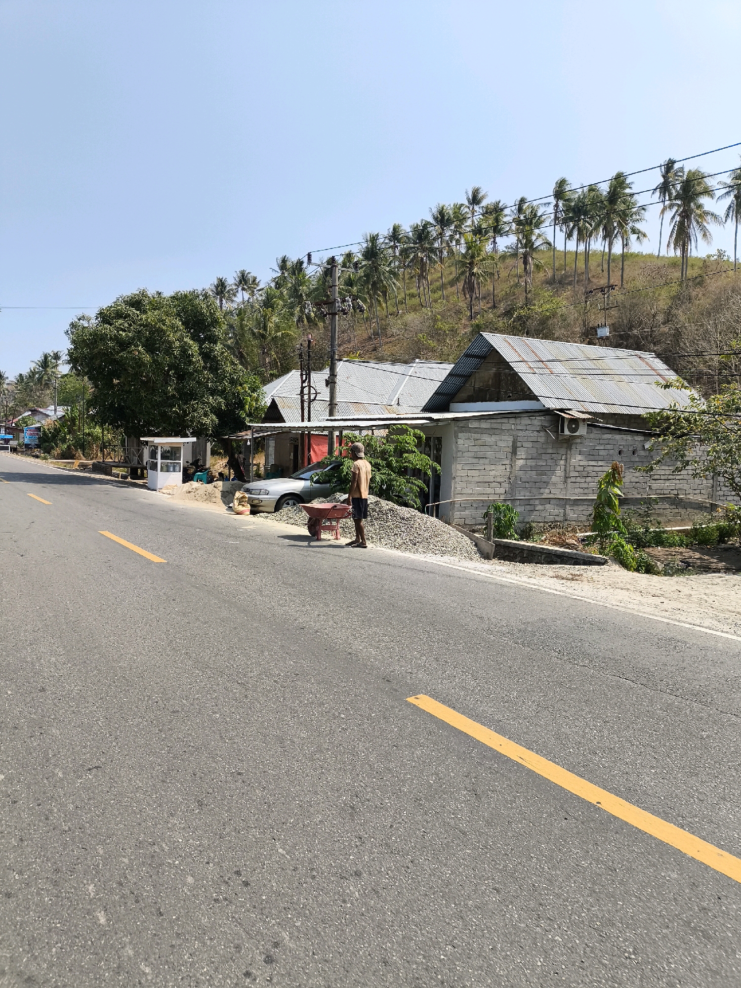 The image depicts a roadside scene in a tropical environment, likely in Indonesia, given the presence of palm trees. A paved road with a yellow dividing line occupies the foreground. To the right of the road, there are several buildings with corrugated metal roofs. One building appears to be constructed of grey bricks, and an air conditioning unit is visible on its exterior wall. In front of the buildings, there is a pile of gravel, a wheelbarrow, and a man wearing a light-colored t-shirt and dark shorts. The man is standing next to the wheelbarrow and the pile of gravel. Further down the road, a silver car is parked. Behind the buildings, a hill is covered with dry grass and lined with numerous palm trees, suggesting a hilly terrain. The sky is clear and blue, indicating a sunny day. There is a small white kiosk or stand on the left side of the road, near a large green tree. The overall scene conveys a sense of a rural or semi-rural area with ongoing construction or material handling.