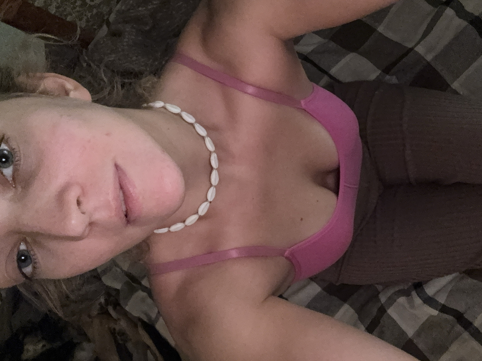 A close-up, angled view of a fair-skinned individual with light blue eyes, light brown hair, and a shell necklace. They are wearing a pink sports bra and brown pants. The background includes a checkered blanket. The location and city of Minsk, Belarus cannot be determined from this image.