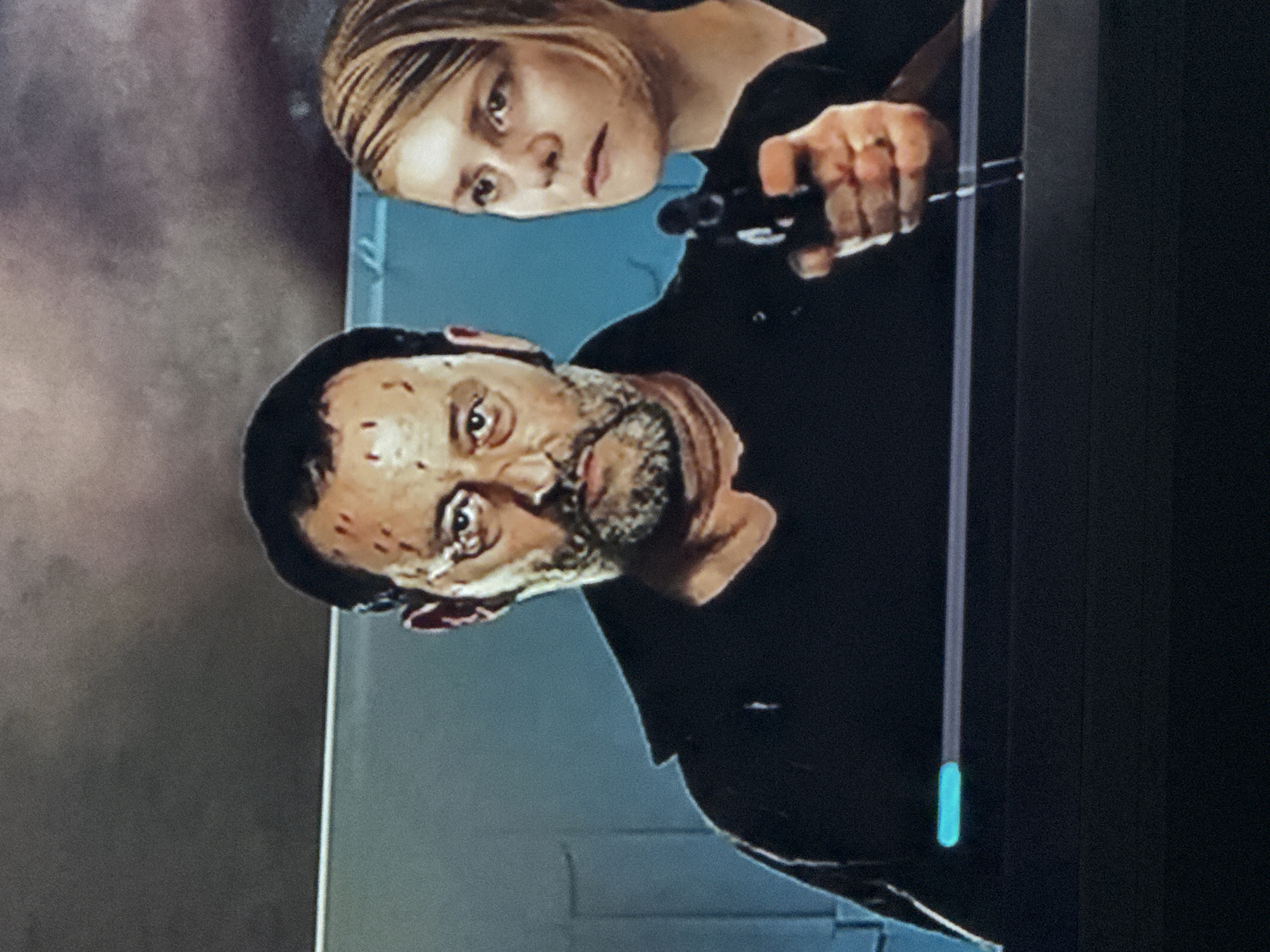 A slightly rotated medium shot from a television screen shows a man and a woman pointing a handgun. The man is in the lower left of the screen and the woman is in the upper center. The man is Caucasian with dark hair, a graying beard, and reddish marks on his face. He is wearing a dark shirt. The woman is light-skinned with blond hair. She is also wearing a dark shirt. Both are holding a handgun. The background behind the man is a bluish-gray wall with horizontal indentations. The context of the television show or the specific location in Mytishchi, Russia, is not ascertainable from the given image.