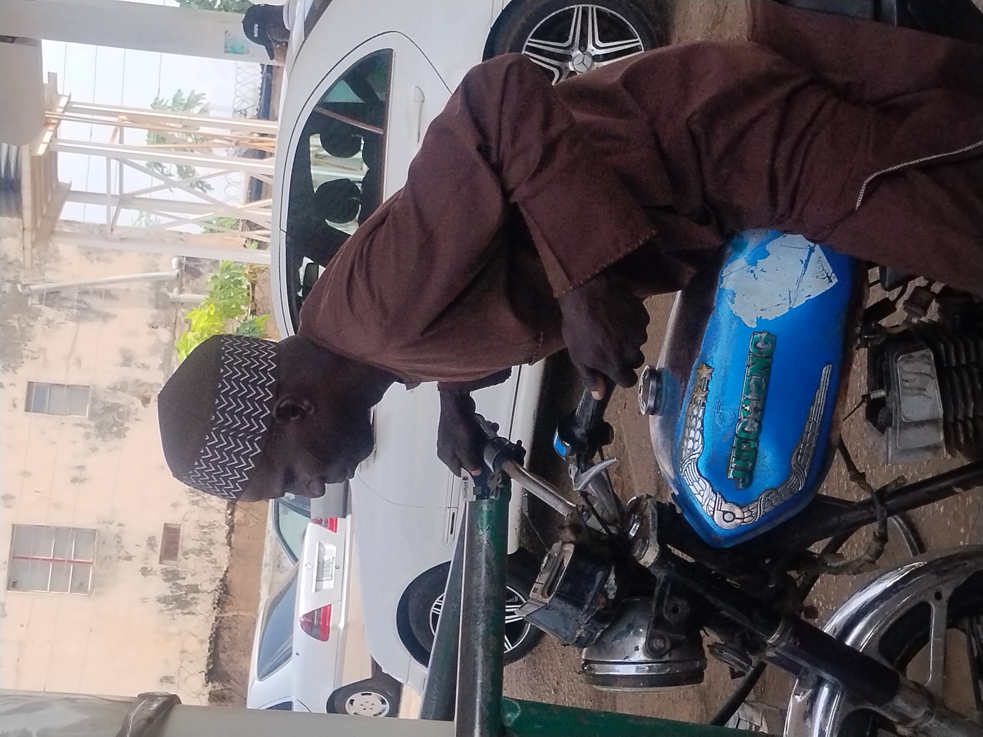 A person dressed in a brown robe and a patterned cap is seen leaning over a blue motorcycle. The person's hands are on the handlebars, suggesting they are either starting the engine or preparing to ride. The motorcycle has a blue fuel tank with decorative silver accents and some peeling paint. The scene appears to be outdoors, with a white car partially visible in the background. The surroundings include a building with windows and some vegetation. The overall lighting suggests it is daytime. The image seems to be taken in Jos, Nigeria, which is suggested by the location context provided.