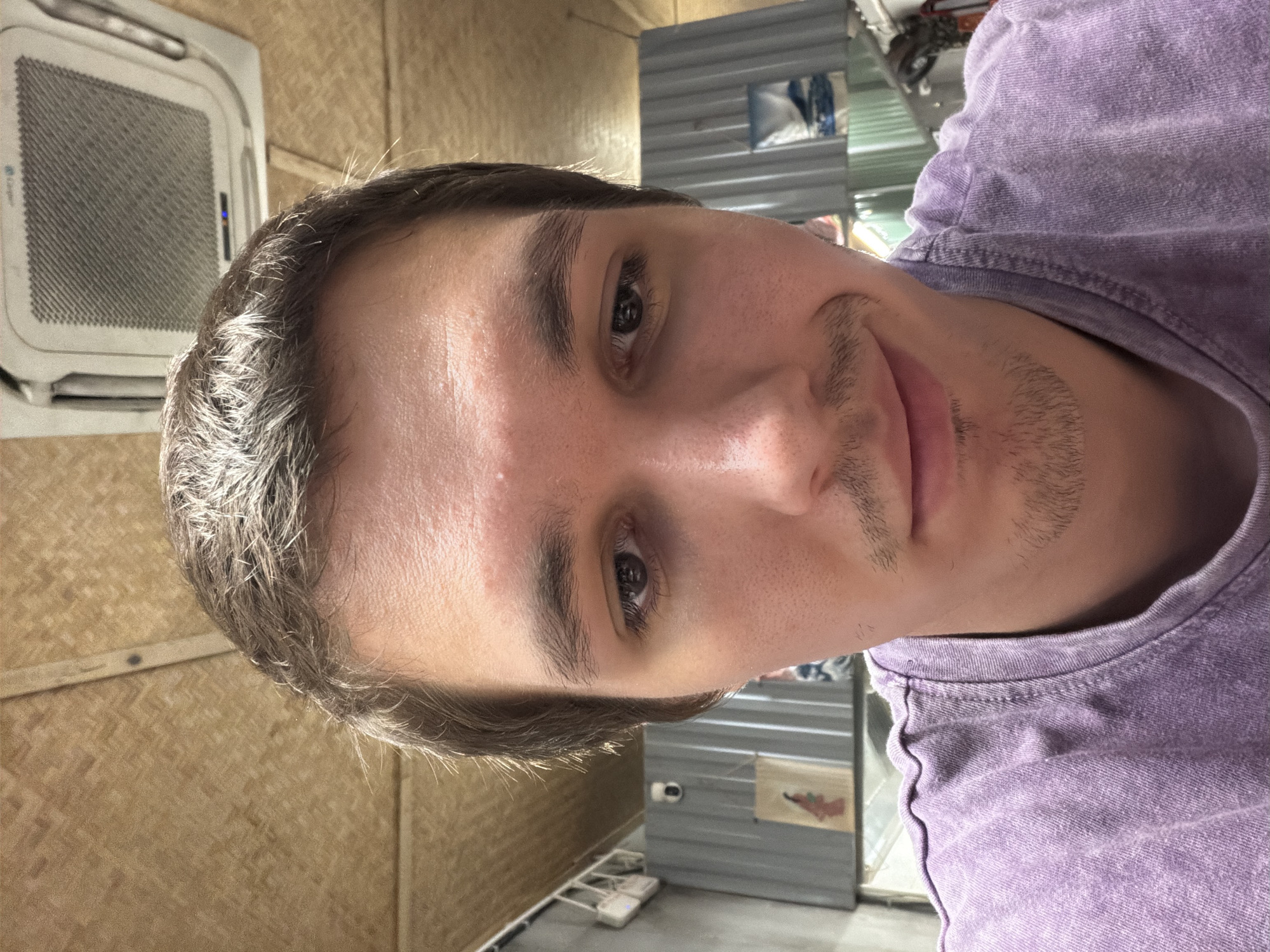 A close-up, rotated portrait shows a young adult male indoors, likely in Đà Nẵng, Vietnam. He has light brown eyes, fair skin, and dark hair with some lighter or graying strands, particularly noticeable where light hits it. He sports light stubble around his mouth and chin. The man looks directly at the camera with a neutral to slightly content expression and is wearing a textured, light purple t-shirt.
The background features distinct elements. To the left of the subject, a wall covered in woven bamboo or rattan matting is visible. Above it, a white, ceiling-mounted LG brand air conditioning unit with a blue indicator light is present. To the right, the background is composed of corrugated metal sheeting, suggesting a partition or wall, with some indistinct items on shelves and several white electronic devices on a low surface. The lighting is bright, indicating a well-lit indoor environment. The "LG" logo is clearly visible on the air conditioning unit.