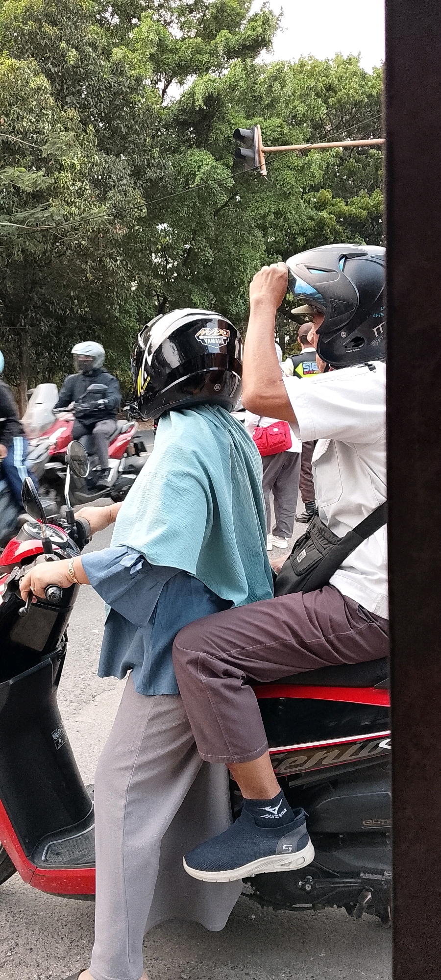 In Depok, Indonesia, two individuals are seated on a red and black motorcycle, captured from a side-view perspective. The person in the foreground, operating the motorcycle, wears a light blue outer garment, gray pants, and dark blue sneakers with white soles and a visible "Mizuno" logo. They have a gold-colored bracelet on their left wrist. Behind them, the passenger wears a white shirt and dark brown pants, along with a black helmet with blue accents and a black crossbody bag. The passenger's right hand is raised, holding the helmet. Both individuals are wearing helmets for safety. The scene appears to be on a street, with trees and a traffic light visible in the background, suggesting an urban or suburban environment. Other motorcycles and people can be seen in the distance, indicating a moderately busy street. The image is framed by a dark vertical element on the right, possibly a window frame or a building edge.