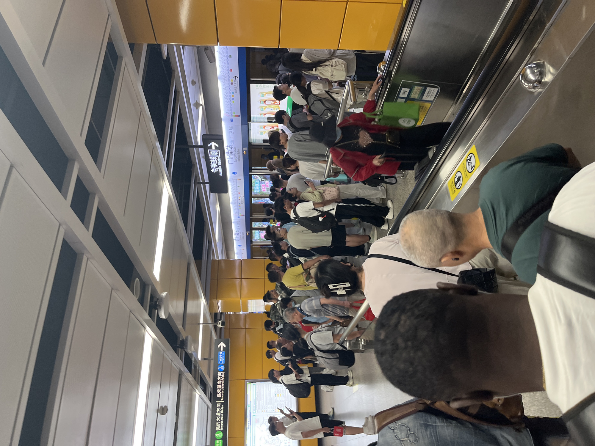 The image captures a scene inside a subway station in Panyu District, China, likely during a busy period. A crowd of people, varying in age and ethnicity, are gathered on an escalator and in the platform area, indicating they are waiting for or disembarking from a train. The station features modern architecture with bright lighting and yellow-colored accent walls. Signage in both Chinese and English is visible, indicating directions to various destinations and station services. Some individuals are looking at their phones, while others are engaged in conversation or observing their surroundings. The overall atmosphere appears to be one of organized movement and transit.