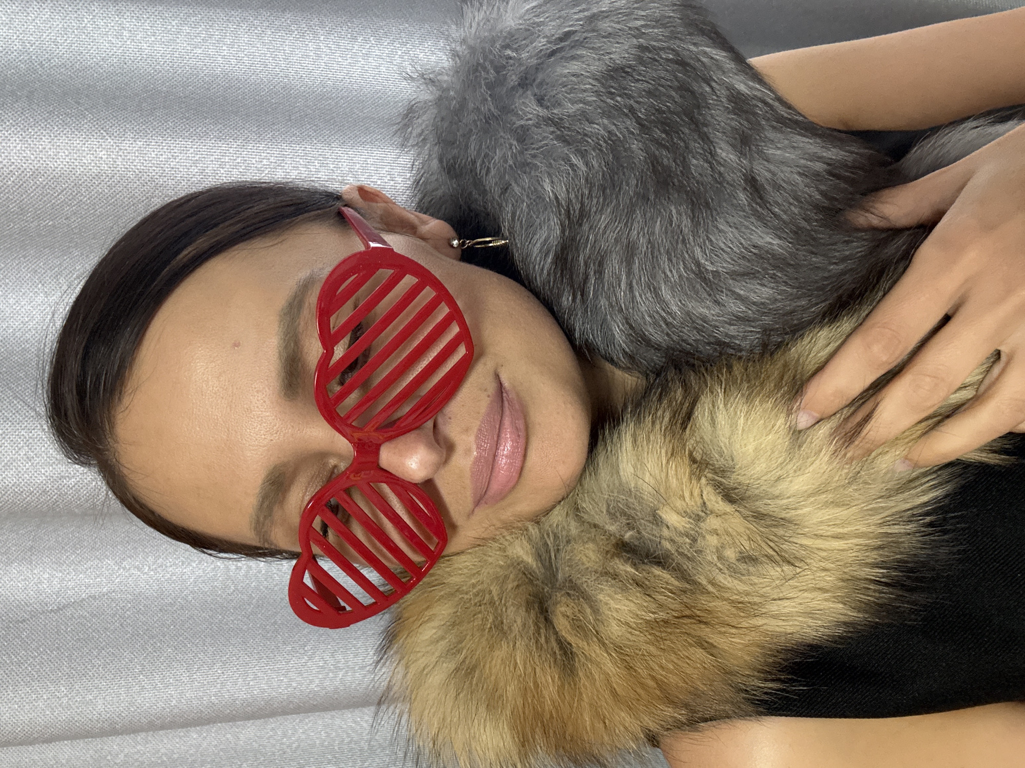 A close-up, eye-level shot features a person, likely a woman, lying on their side on a light gray, subtly textured surface, possibly a bed or sofa. They are wearing distinctive red, heart-shaped shutter shades. A luxurious, two-toned faux fur garment, comprising a grayish-silver section near the head and a brownish-gold part extending towards the chest, drapes around them. The person has dark hair pulled back and exhibits a subtle, relaxed smile with glossy lips. A small, delicate earring is visible in their left ear, and one hand gently rests on the fur. The image is brightly and evenly lit, indicating an indoor setting. No text is visible. The scene is set in Chaikovsky, Russia.