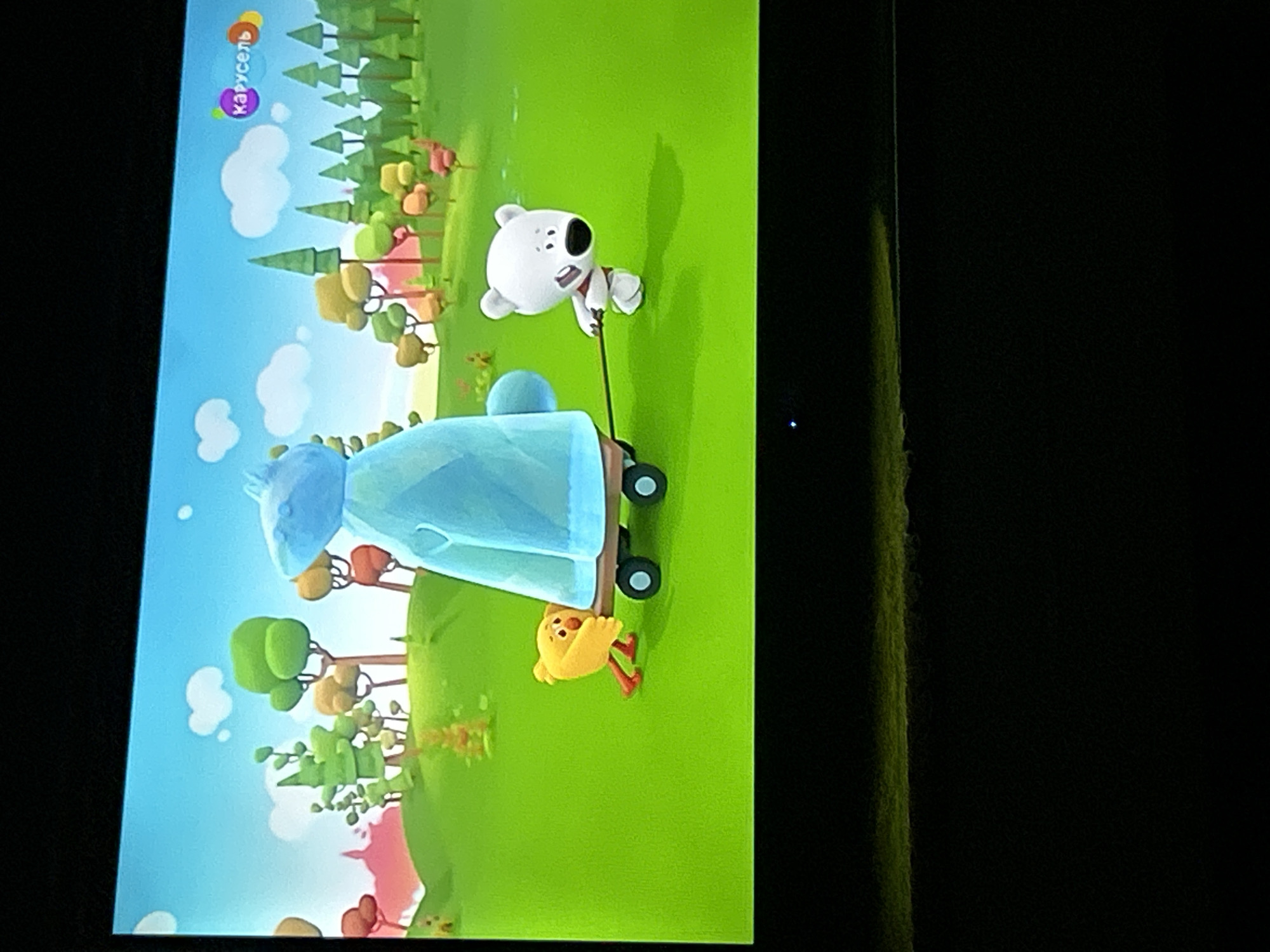A television screen displays a cartoon scene with a bright green grassy field under a blue sky with scattered white clouds. In the foreground, a small yellow cartoon chick is pushing a blue object on a two-wheeled cart. To the right of the cart, a white cartoon bear is pulling the cart with a dark rope. The bear is wearing a red ribbon around its neck. In the background, there are stylized trees with green and orange foliage, and some pink hills. In the upper right corner of the screen, a logo with the text "Карусель" is visible. The image is captured from an angle, with the bottom portion of the screen showing a black area, indicating it's a photograph of a television. The scene appears to be from a children's animated show. There is no specific information to suggest the location is Gelendzhik, Russia.