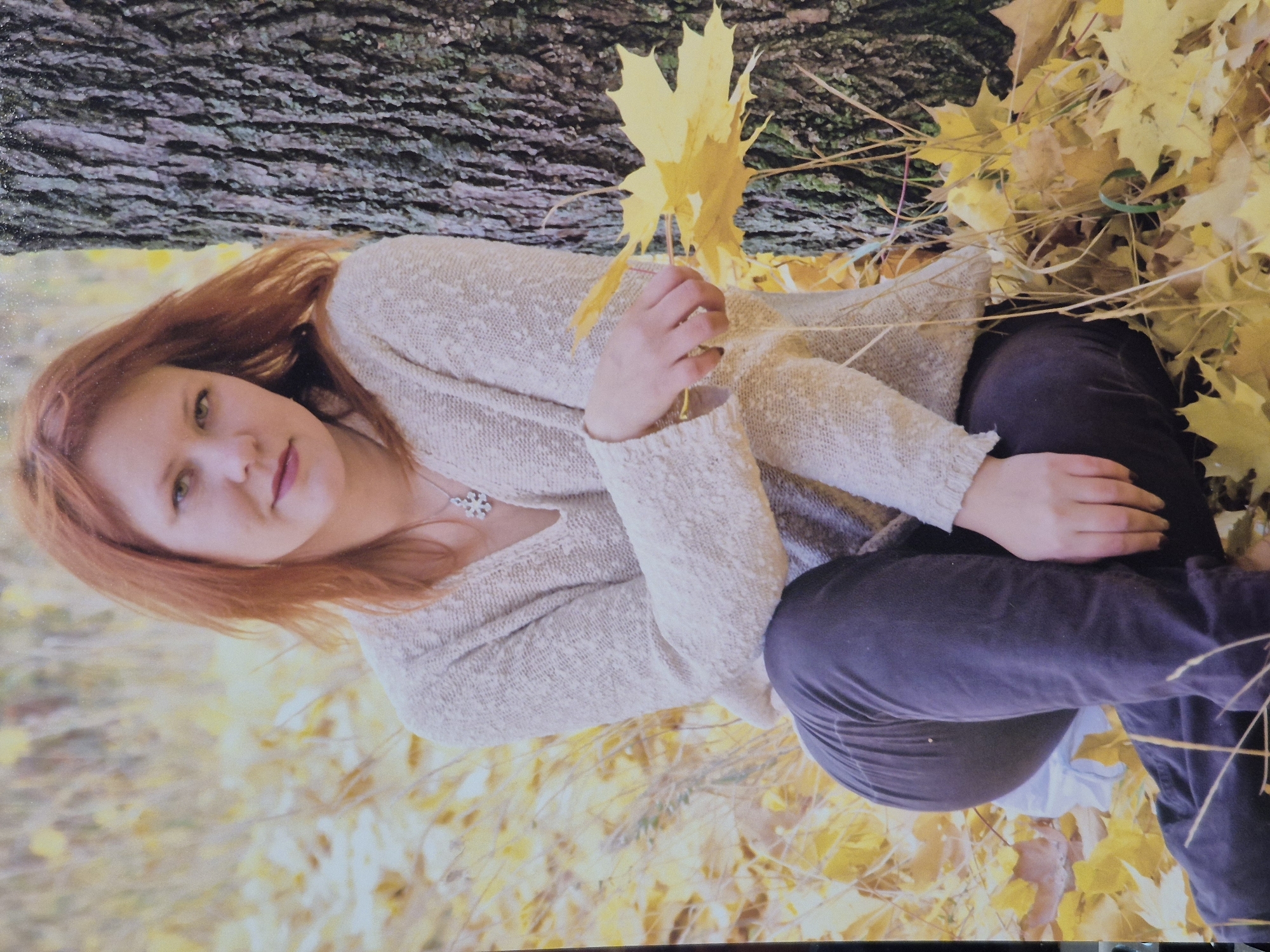 A woman with red hair sits on the ground in an outdoor setting, leaning against a tree. She is wearing a light-colored, textured sweater and dark pants. Her arms are crossed in front of her, and she is holding a few yellow leaves in her right hand. The ground around her is covered with fallen yellow leaves, suggesting an autumn season. The tree trunk is rough and textured. The background is softly blurred, with a mix of yellow and brown tones, further indicating an autumnal environment. The scene appears to be a portrait-style photograph, possibly taken in a park or wooded area within the city of Schwäbisch Gmünd, Germany.