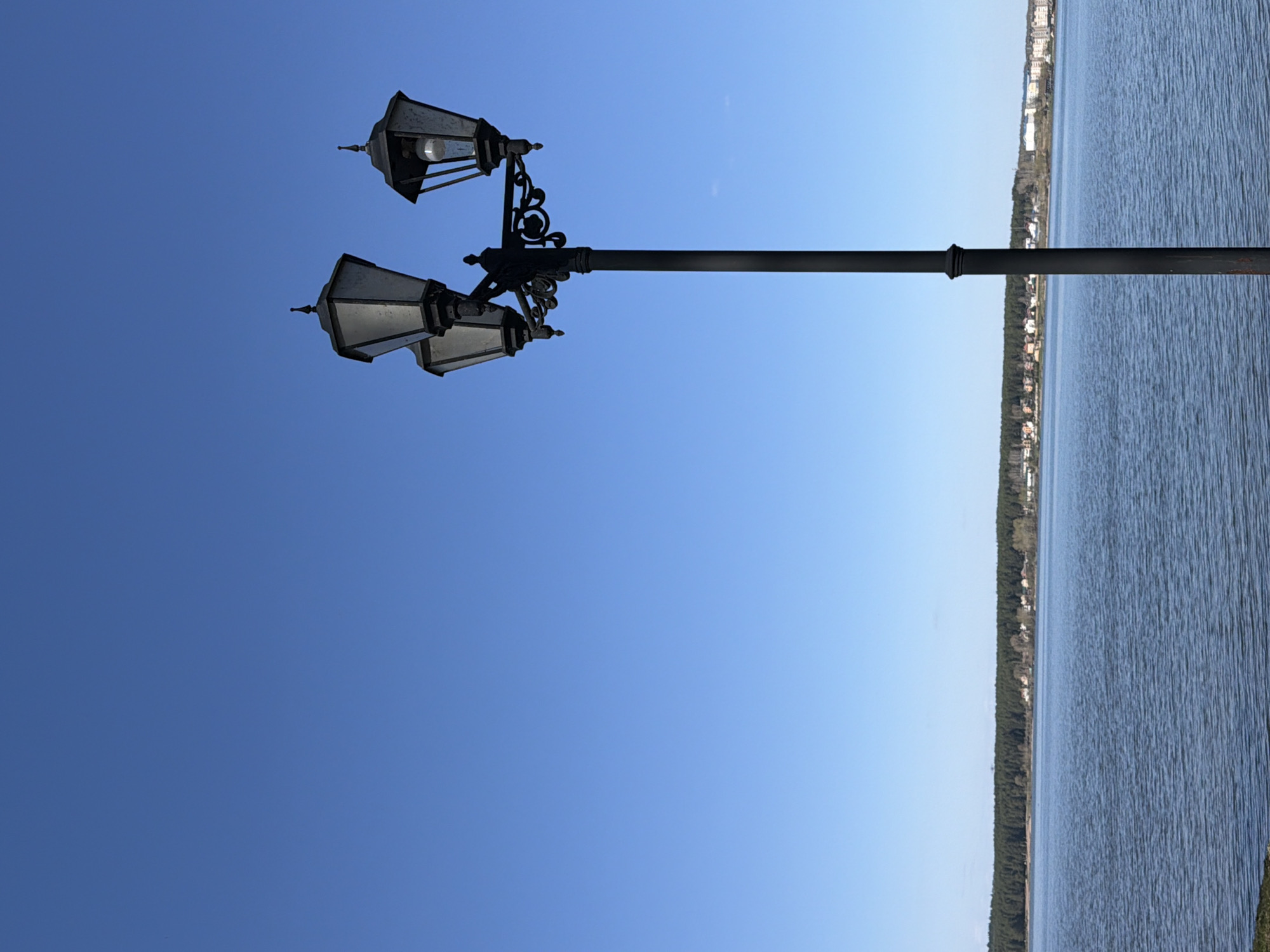 The photograph captures a street lamp in Votkinsk, Russia, with three ornate lanterns attached to a black, horizontal pole. The lanterns are a dark color and appear to be made of metal and glass. The top lantern has a bulb visible within, while the lower two are obscured. The pole extends toward the right side of the frame, where a body of water stretches into the distance, lined with trees and buildings along its shore. The sky overhead is a clear, bright blue. The image is taken from a low angle, with the horizon oriented vertically.