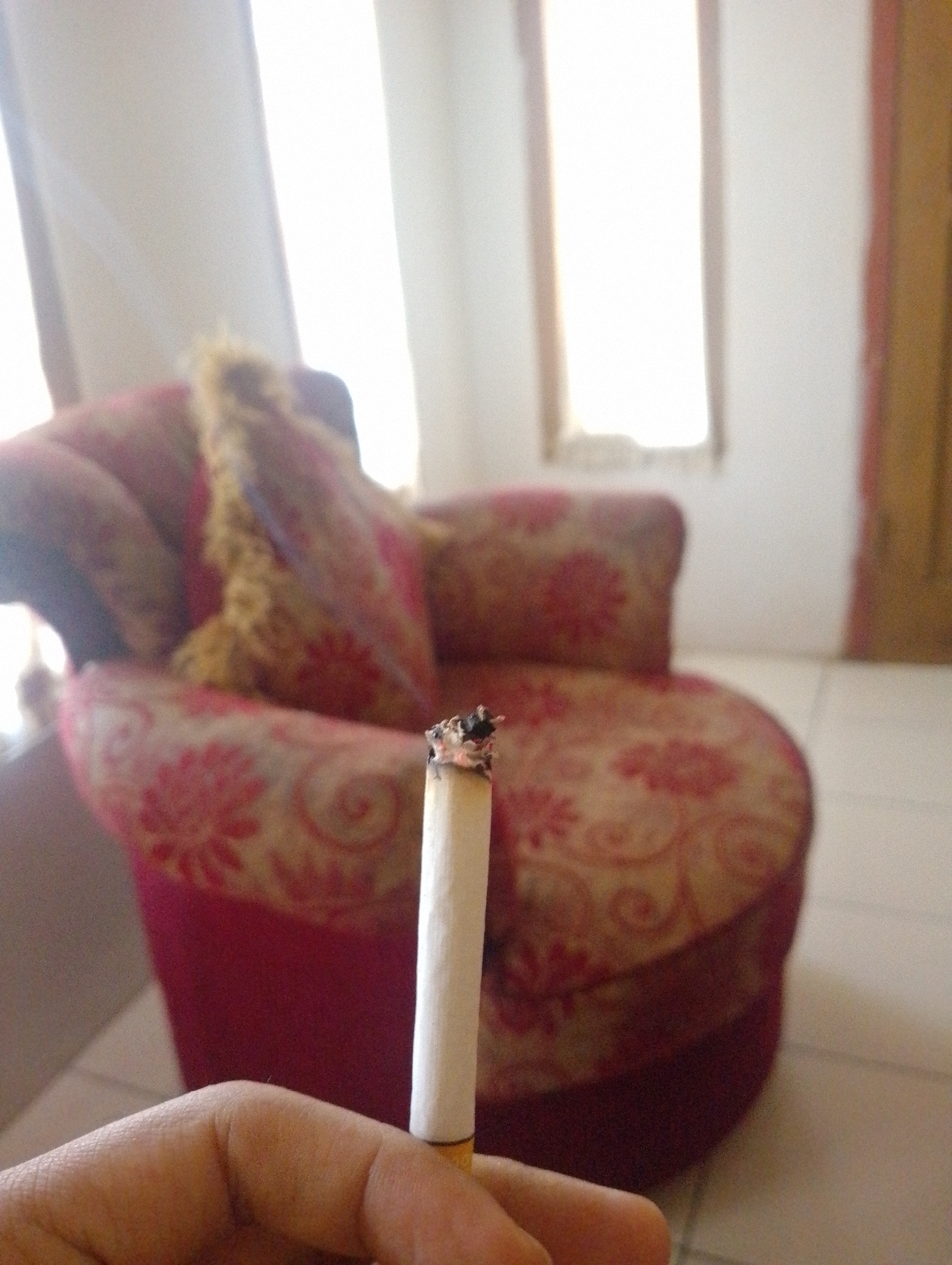 The image depicts a close-up of a hand holding a lit cigarette in the foreground. The cigarette is white with a brown filter, and ash is visible at its tip. In the background, a red upholstered armchair with a floral pattern is visible. The armchair appears to be in a room with natural light streaming in from what looks like a window covered by sheer curtains. The overall setting suggests an indoor environment, possibly a living room or lounge. No specific landmarks of Sukamanah, Indonesia, are identifiable in the image.