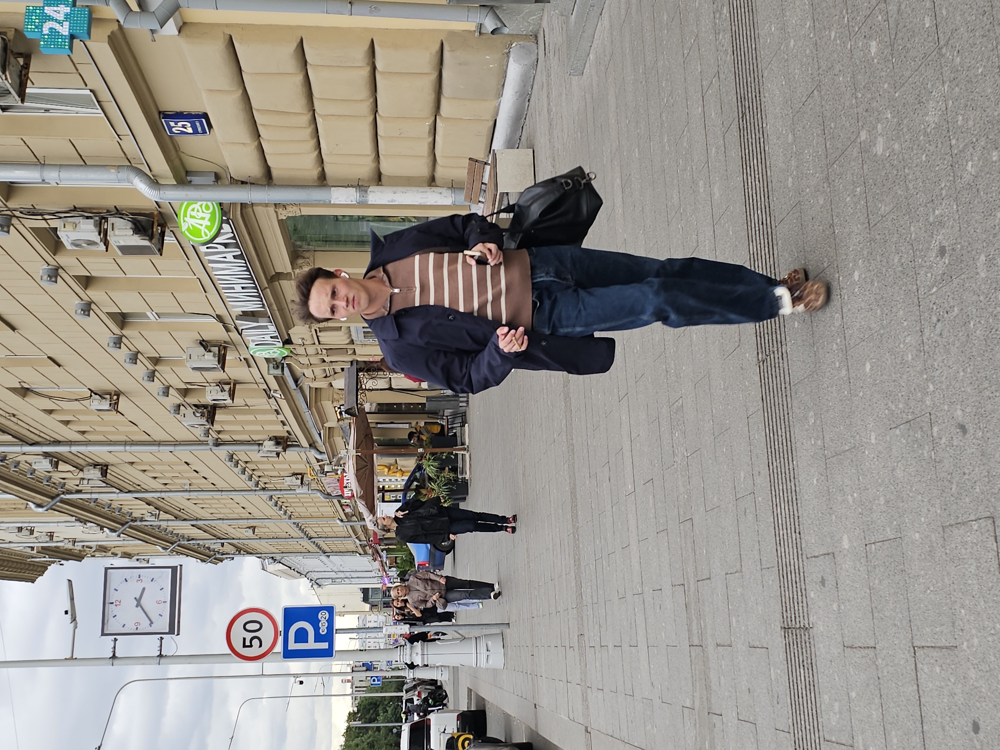 The image depicts a street scene in Moscow, Russia, likely on a cloudy day. In the foreground, a young Caucasian man with light brown hair is walking towards the viewer on a grey, paved sidewalk. He is wearing a dark blue trench coat over a striped brown and white quarter-zip sweater, dark blue jeans, and light brown shoes. He has white wireless earbuds in his ears and is holding a smartphone in his right hand and a dark leather bag in his left.
To the left of the man, a large square clock with Roman numerals is mounted on a pole. Below the clock, a circular red sign with "50" in white text indicates a speed limit, and a blue rectangular sign with a white "P" and smaller numbers "10, 15, 20" likely indicates parking restrictions. Further down the sidewalk, other pedestrians are visible.
The background features a multi-story building with a pale yellow facade. Several air conditioning units are mounted on the building. A street sign with the number "25" is visible, along with a blue sign with a green plus symbol and the number "24" in a bright blue pixelated font, suggesting a 24-hour establishment. A striped awning from a cafe or shop extends from the building. Cars are parked along the street. The sidewalk is made of large, light grey rectangular paving stones with a textured strip for tactile guidance for the visually impaired.