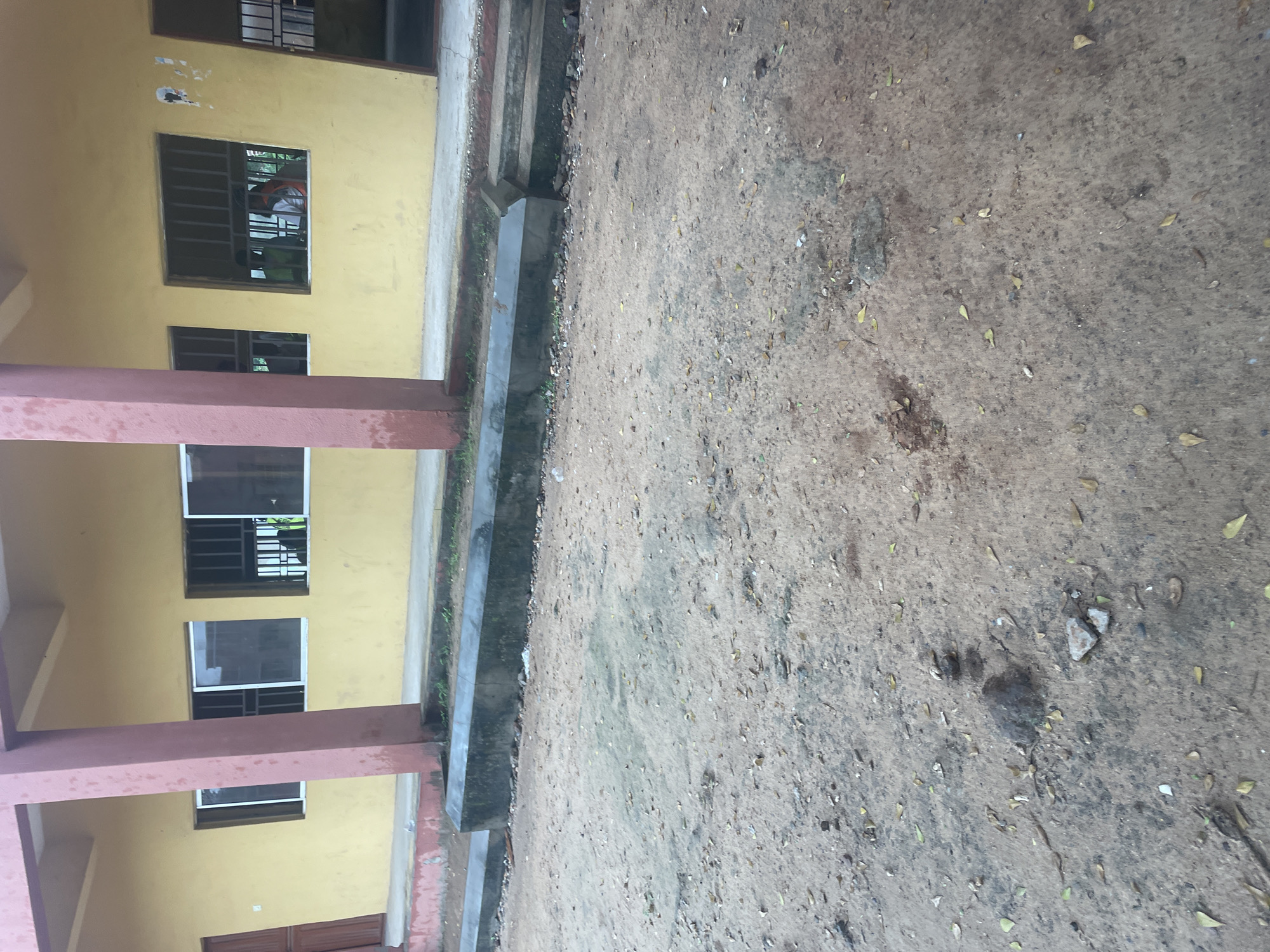 This is an outdoor, eye-level, medium shot of a building facade and the ground in front of it. The building is painted yellow and has several windows with metal bars. The ground is covered in sand, dirt, and scattered dry leaves. A concrete barrier runs along the base of the building. In one of the windows, two people are visible inside, appearing to be students, wearing vests. The location is identified as a school in Lokoja, Nigeria.