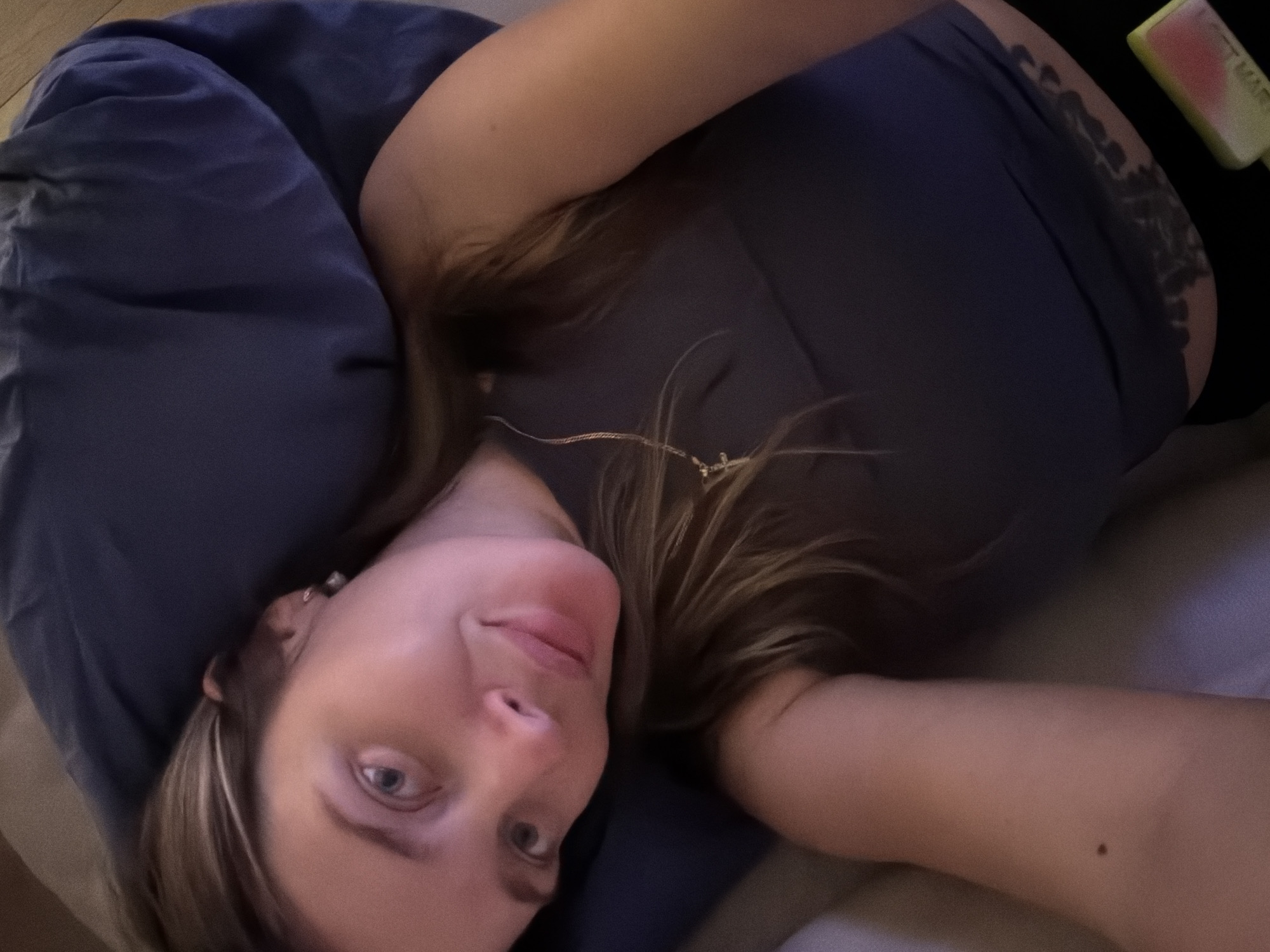 A woman is lying on her side, resting her head on a navy blue pillow. She is wearing a dark grey tank top. A thin gold chain necklace with a small cross pendant is visible around her neck. Her hair is long and brown, falling around her face and shoulders. A tattoo is visible on her lower abdomen, extending to her right hip. The background appears to be a bed. There is no indication of the city of Moscow, Russia within the image.