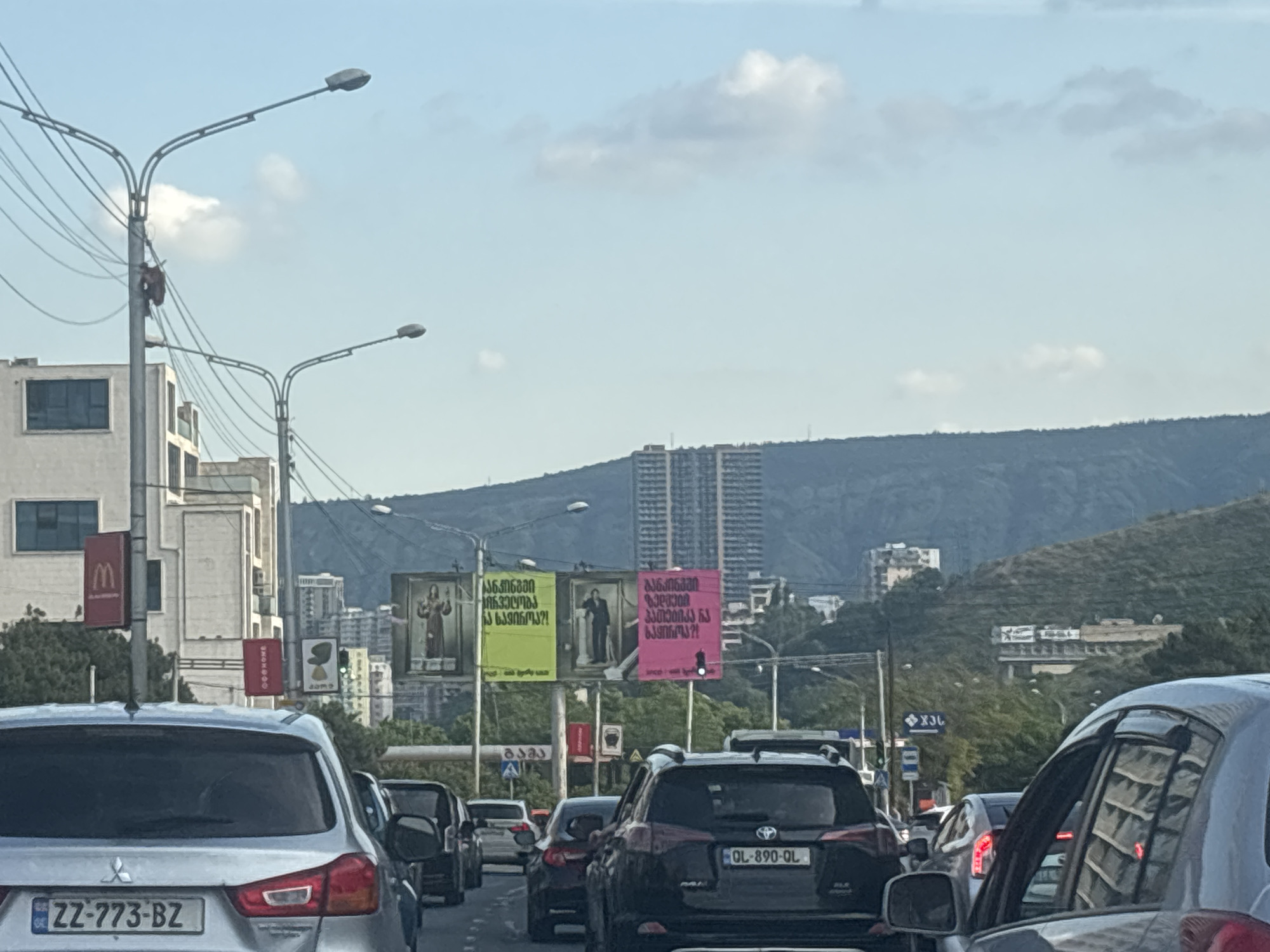 The image captures a traffic scene in Tbilisi, Georgia, characterized by a line of cars on a wide road. In the background, a densely vegetated hillside rises, dotted with buildings of various sizes, including a prominent tall, modern structure. Along the roadside, there are billboards with Georgian text and images. One billboard features a pink background with text and an image of a person in formal attire. Another billboard has a yellow background with similar text and an image of a person in religious garments. A McDonald's sign is visible on a pole, suggesting proximity to commercial establishments. Streetlights and power lines are overhead, indicating urban infrastructure. The sky is partly cloudy, with a clear blue expanse. The cars are stopped or moving slowly, typical of urban traffic. The license plate of the silver car in the foreground reads "ZZ-773-BZ", and the black SUV behind it has a license plate "QL-890-QL". The overall impression is one of a busy urban environment with a mix of modern and older architecture.