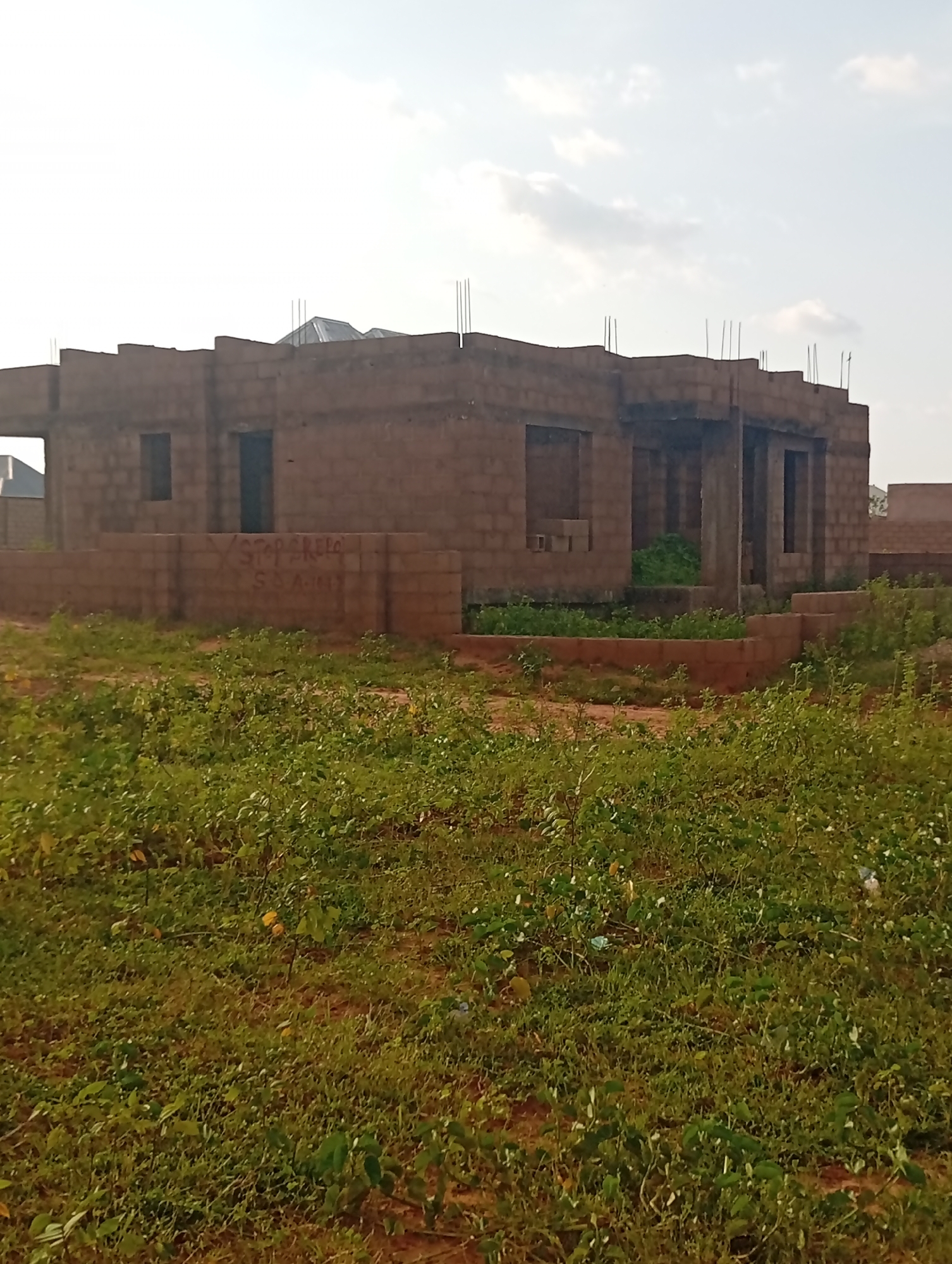 The image depicts an unfinished building constructed from reddish-brown bricks, located in the city of Tumfure, Nigeria. The structure appears to be a single-story dwelling with open doorways and window frames. Several metal rebar rods protrude from the top of the walls, indicating that further construction is planned. The building is surrounded by overgrown green vegetation and a patch of dry earth. A low brick wall, possibly part of the foundation or an outer boundary, is visible in front of the building. Graffiti is present on this low wall, with text that is partially obscured but includes the words "STOP GREE" and "S.S.A." The sky above is light and hazy, suggesting it is daytime, possibly late afternoon. No people are visible in the image.