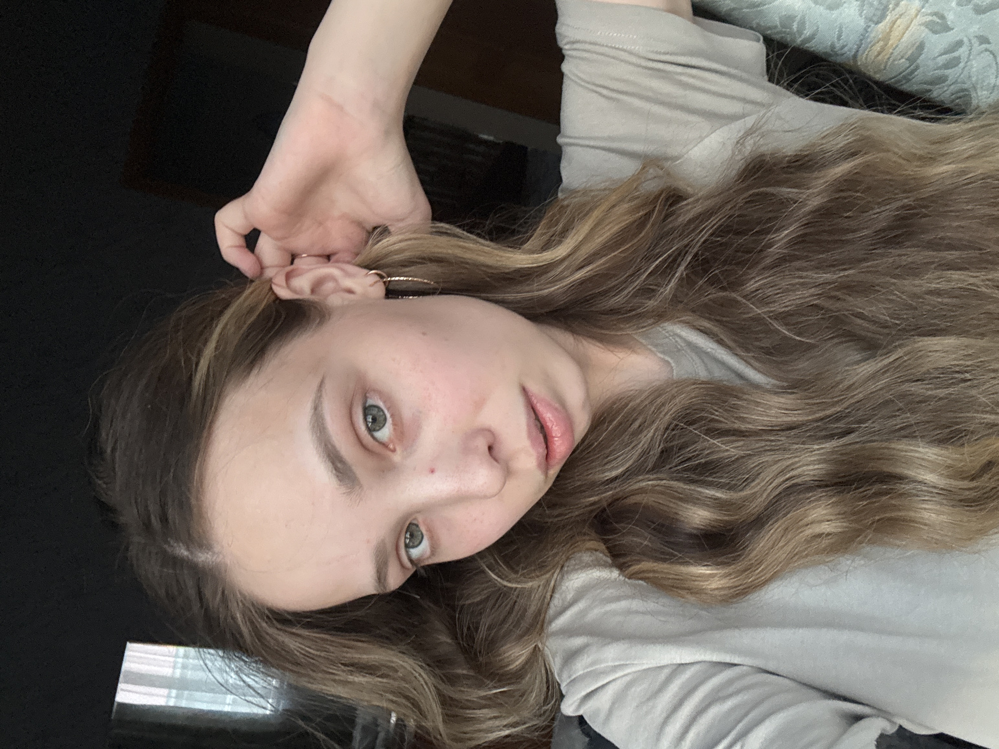 A young woman with long, wavy, light brown hair is posing for a selfie indoors. She is wearing a light gray t-shirt and gold hoop earrings. Her left hand is near her ear. The background includes a dark, possibly black, surface and a light-colored patterned surface. The location in the city of Прикамский, Russia is not determinable from the image.