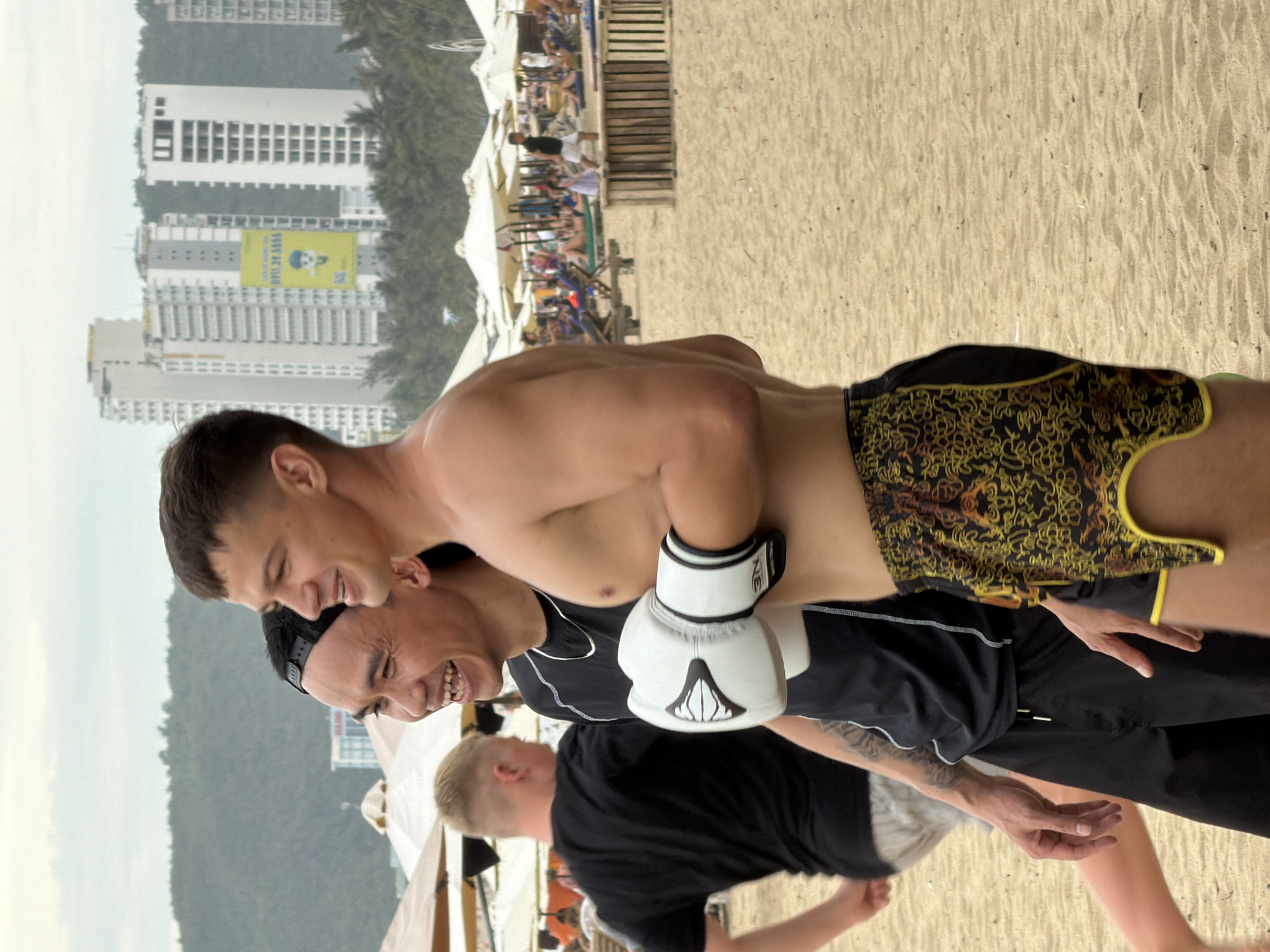 Two men are standing on a sandy beach, facing right and smiling. The man in the foreground is wearing black and yellow patterned shorts and is shirtless. He is holding white boxing gloves. The man behind him is wearing a black t-shirt and black shorts, and also has boxing gloves on. In the background, there are beach umbrellas, lounge chairs, and several tall buildings, suggesting an urban beach setting in Nha Trang, Vietnam. The sky is overcast.