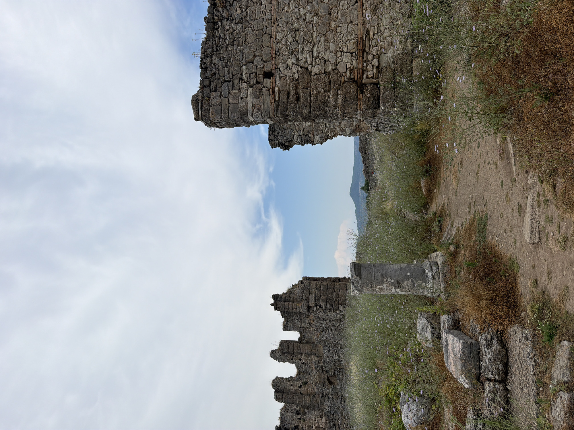 The image captures a view of ancient ruins in Serik, Turkey, likely within the archaeological site of Aspendos. In the foreground, a dirt path winds through overgrown vegetation, dotted with small purple wildflowers. Scattered on the ground are fragments of stone, including a large, rectangular block resting on smaller stones, and a broken pillar. To the right, a section of a large, weathered stone wall dominates the frame, with visible layers of different colored stone and brickwork. To the left, the jagged ruins of another stone structure rise against a backdrop of a hazy sky with scattered clouds. In the distance, rolling hills are visible, suggesting a natural landscape surrounding the ancient site. The overall impression is one of a historical site being reclaimed by nature.