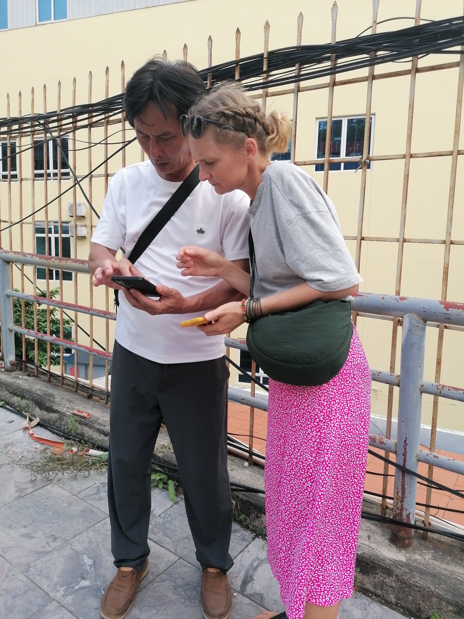 Two individuals, a man and a woman, are standing outdoors, intently looking at and interacting with their smartphones. The scene appears to be an elevated walkway or platform, possibly a rooftop, in Hong Gai Ward, Vietnam.
The man, who appears to be Asian, is on the left, wearing a white short-sleeved t-shirt with "allo" or "allô" visible on the chest, dark grey or black pants, and brown loafers. He has dark hair and a black cross-body bag slung over his shoulder. He holds a black smartphone in both hands, his fingers poised over the screen.
The woman, appearing Caucasian, is on the right, with blonde hair styled in braids, and sunglasses pushed up onto her head. She wears a loose-fitting grey t-shirt, a long pink skirt with a white patterned design, and a dark green cross-body bag. Her left wrist is adorned with multiple bracelets. She holds a yellow smartphone in her right hand, her gaze directed towards its screen, while also seeming to observe the man's phone.
In the background, a light yellow building with multiple windows and air conditioning units is visible. A rusty metal railing with numerous black electrical wires running across it separates the subjects from the building. The ground is made of concrete paving, with some debris and sparse vegetation visible near the base of the railing. The lighting suggests a bright, possibly overcast, daytime. Both individuals appear focused and engaged with their devices, possibly navigating or sharing information.