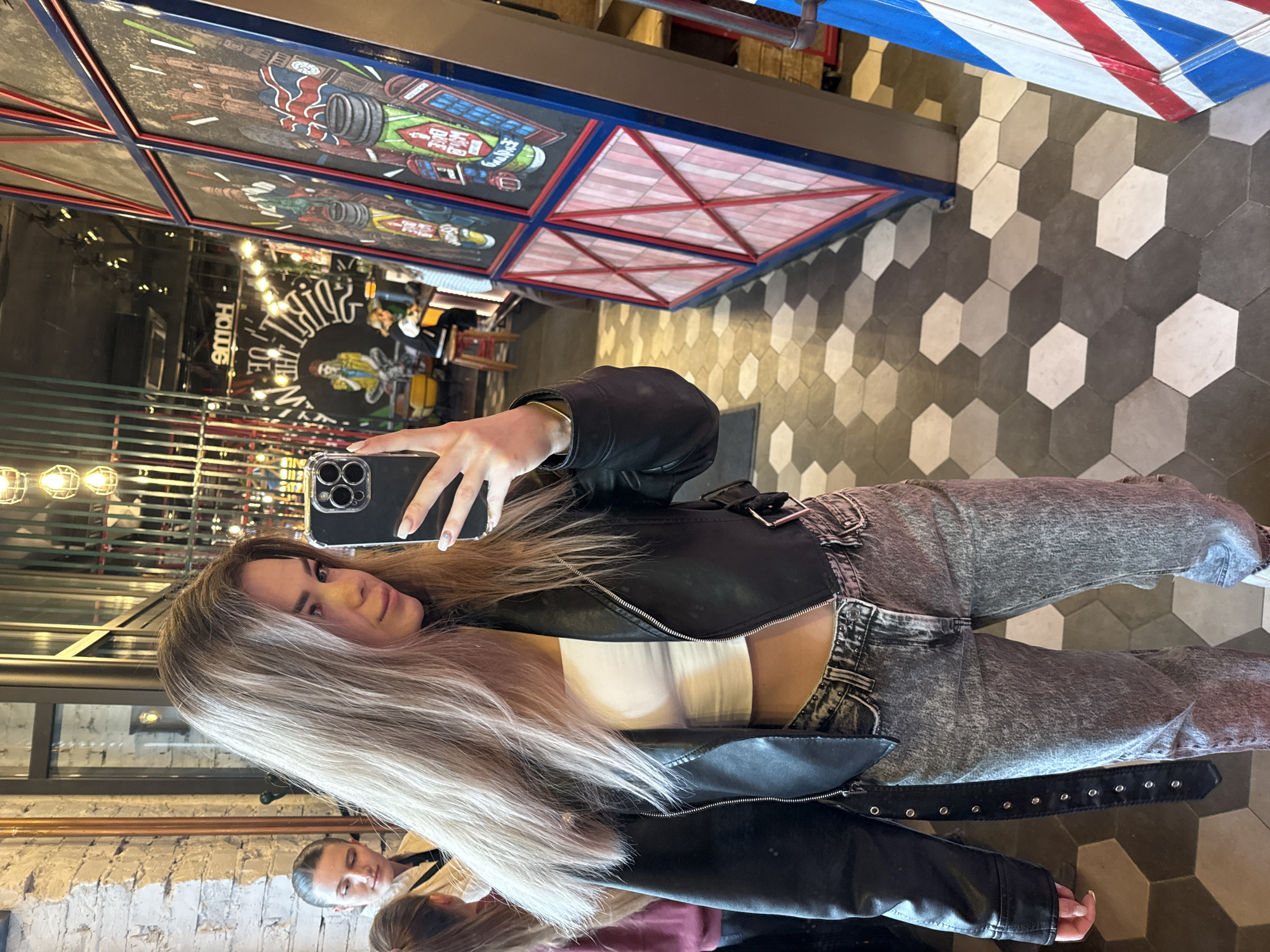 Here is a detailed description of the image:
The image is a selfie taken in what appears to be a barber shop or a similar establishment with a retro or vintage design. The main subject is a woman with long blonde hair, wearing a black leather jacket over a white top and acid wash jeans. She is holding a smartphone, presumably taking the selfie in front of a mirror.
The background shows details of the location:
- The floor is tiled with a hexagon pattern in grayscale colors.
- Above, there is a structure with framed artworks or designs, featuring vintage-style illustrations and geometric patterns, with some sections painted in red and white.
- The reflection in the mirror shows interior details like lighting fixtures and other people, indicating that it is a commercial space.
The lighting seems to be artificial, and the overall style suggests the establishment is aiming for a classic or nostalgic aesthetic. The scene is urban, implying it's in a city, but the specific location is not identifiable from the image.
