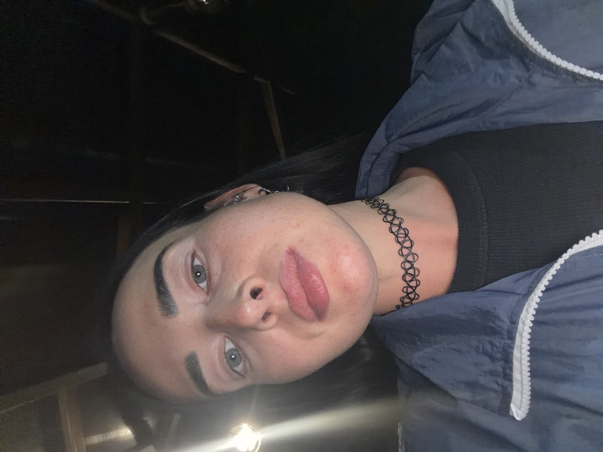 A low-angle, medium shot shows a young adult with dark hair, fair skin, and light blue eyes looking at the camera. The subject is wearing a navy blue windbreaker with a white zipper and a black ribbed shirt underneath. A black choker necklace is visible around their neck. The background is dark and appears to be an indoor setting with some lighting visible on the left side. The image is focused on the subject's face and upper body. The location is not depicted in the image.