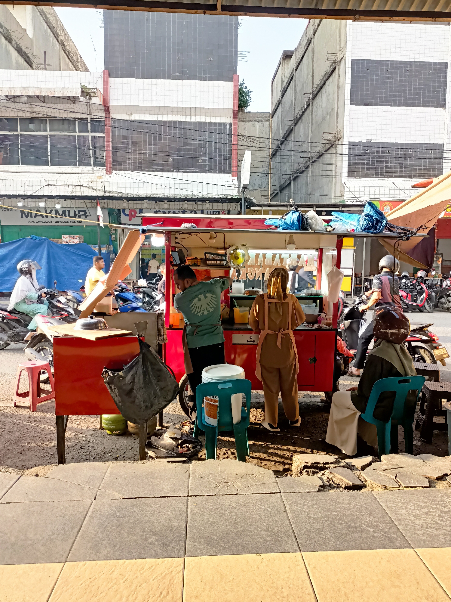 The image depicts a street scene in Bandarbireuen, Indonesia, likely a market or commercial area. In the foreground, a red food cart is set up on a sidewalk. A man in a green shirt is working behind the cart, seemingly preparing food. A woman in a brown hijab and apron is standing in front of the cart. Several customers are visible. One person, wearing a helmet, is on a motorcycle to the right, facing away from the camera. Another person, also wearing a helmet and a brown garment, is seated on a blue plastic chair to the right of the food cart, observing. In the background, there are buildings and several motorcycles parked on the street, suggesting a busy urban environment. The sidewalk shows signs of disrepair with cracked tiles. The overall atmosphere appears to be that of a bustling street-side food vendor.