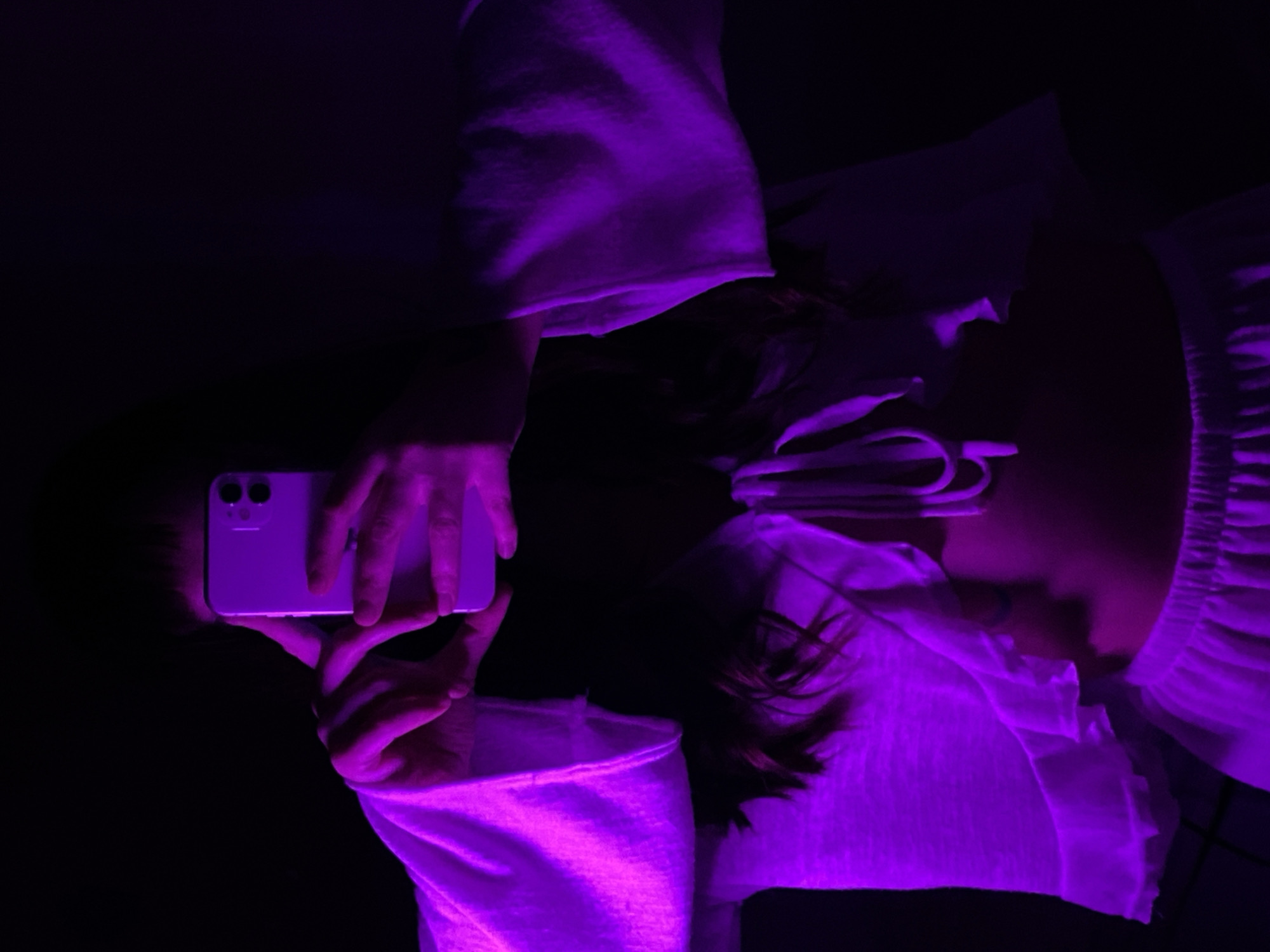 A person lies on their back in a dark environment illuminated by a purple light, holding a white smartphone. The person's face, partially visible, is turned to the side. They wear a white top with ruffled sleeves. Their hair is dark. The setting is nondescript, lacking distinctive features that would suggest a location in Votkinsk, Russia, or elsewhere.