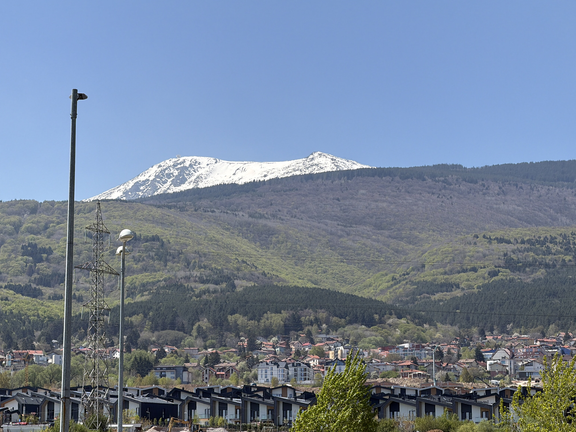 The image displays a panoramic view of a town located at the base of a majestic, snow-capped mountain under a clear, bright blue sky. This setting is identifiable as Sofia, Bulgaria, with the prominent mountain likely being Vitosha.
The mountain dominates the background, its upper reaches still covered in white snow, indicating a late winter or early spring season. Below the snowline, the slopes are densely forested, featuring a mix of darker green coniferous trees and lighter green deciduous trees that are just beginning to leaf out.
In the mid-ground and foreground, a sprawling urban or suburban settlement is visible. The immediate foreground features a row of modern residential buildings, characterized by dark roofs and light-colored walls, some appearing to be recently built or under construction. Beyond these, the town extends with a diverse array of buildings, many with red, orange, and brown tiled roofs, typical of the region.
The time of day appears to be mid-morning to early afternoon, given the bright sunlight and clear shadows. The weather is excellent, with a completely cloudless sky. Infrastructure elements are noticeable, including a tall metal electricity pylon prominently positioned in the left-mid foreground, and several power lines traversing the scene. Two tall utility poles or streetlights are also visible on the left. Fresh green foliage on trees within the town further emphasizes the springtime setting. No visible text is present in the image. The scene captures the contrast between the pristine natural beauty of the mountain and the expanding human settlement below.