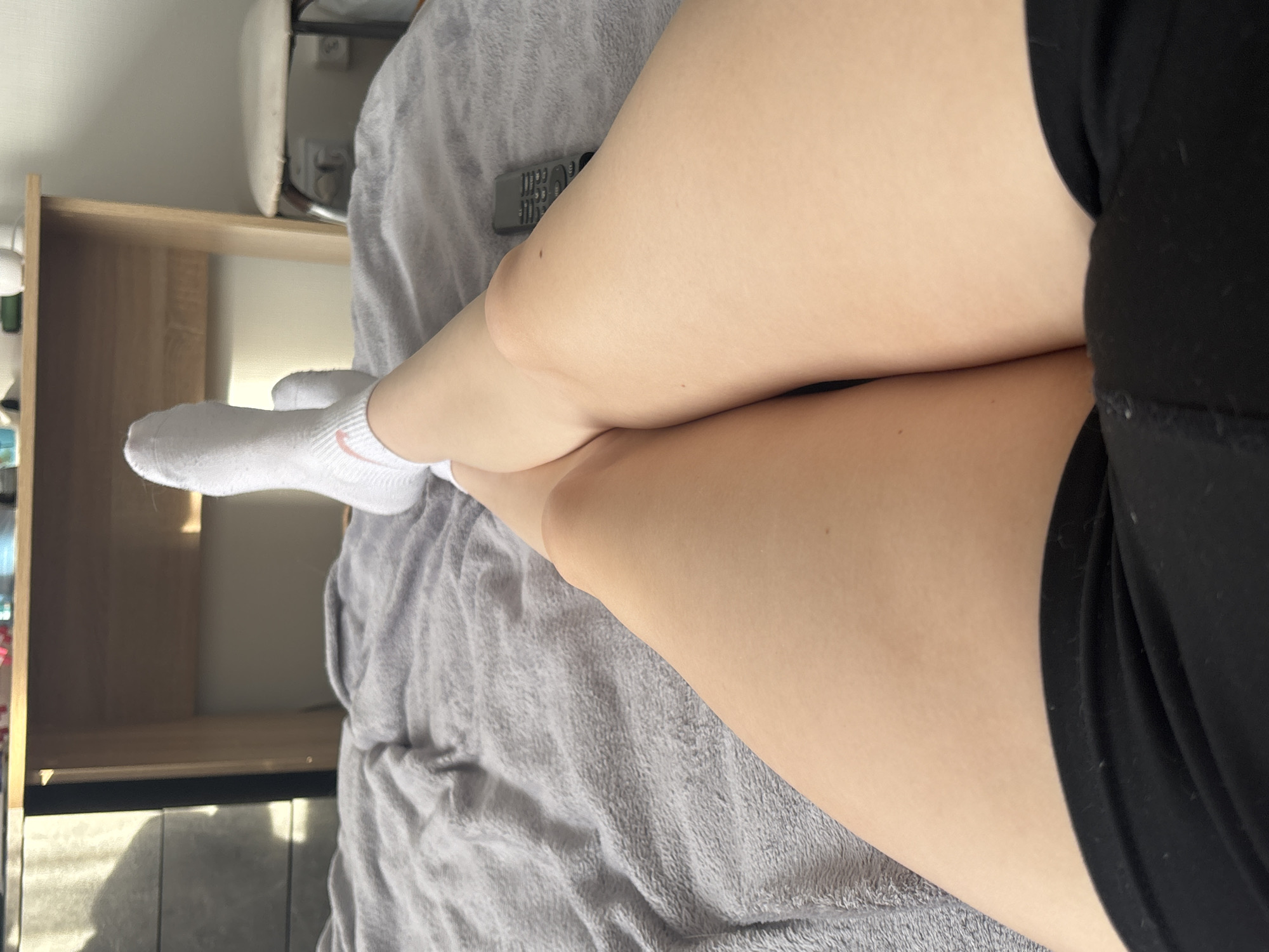 The image displays a close-up view of a person's legs, seen from a high angle as they rest in a relaxed position. The legs, from the upper thigh to the feet, are bare and appear light-skinned. The person is wearing black shorts, partially visible on the right side of the frame. The left foot is adorned with a white athletic sock featuring a distinct pink Nike swoosh logo on the ankle. The other foot is mostly out of view.
The legs are comfortably positioned on a soft, grey, textured blanket or throw that covers most of the underlying surface. A silver-grey remote control is visible on the blanket, situated between the knees, showing its buttons and some indistinct text. In the background, wooden shelving or furniture, possibly a bookcase or cabinet, can be seen. Further back, a white object, which might be a chair back or an ironing board, is partially visible.
The scene is indoors, likely a domestic setting such as a living room or bedroom. Bright, soft lighting, indicative of natural daylight, illuminates the space and casts gentle shadows. The overall impression is one of leisure, suggesting the person is lounging or relaxing, potentially watching television given the presence of the remote control. The image is oriented 90 degrees clockwise from a standard upright view.