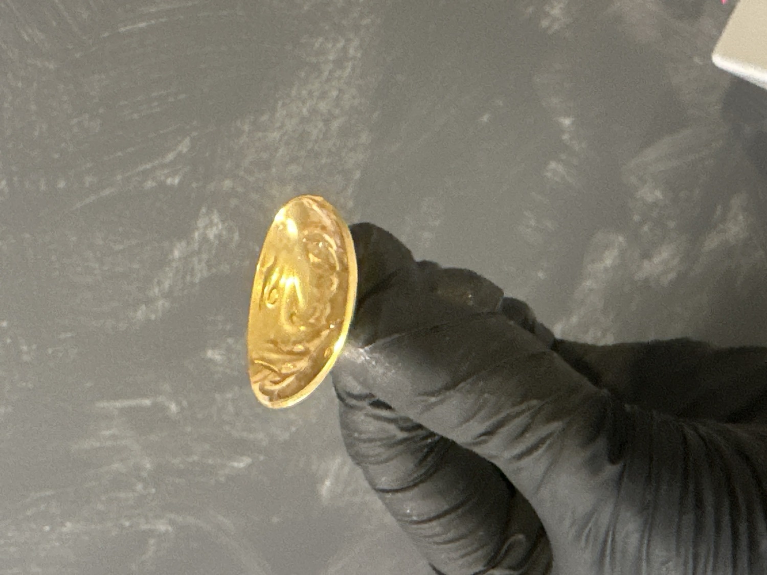 The image captures a close-up of a hand, gloved in dark, possibly black, material, carefully holding a circular, golden-colored object. The object, resembling a coin or medallion, is held between the thumb and index finger, angled to reveal its surface. Intricate, embossed patterns are visible on the golden item, which seem to depict a profile, possibly of a person, or an ornate emblem. Some indistinct shapes that could be partial text or numbers are present within the design, though unreadable. The background is a textured, dark gray surface, consistent with a workbench or table, exhibiting lighter, streaked patterns that suggest wear or a particular finish. The scene portrays the meticulous handling and presentation of a potentially valuable artifact. The protective glove indicates a need for careful examination or preservation. The activity is occurring indoors in Riga, Latvia.
