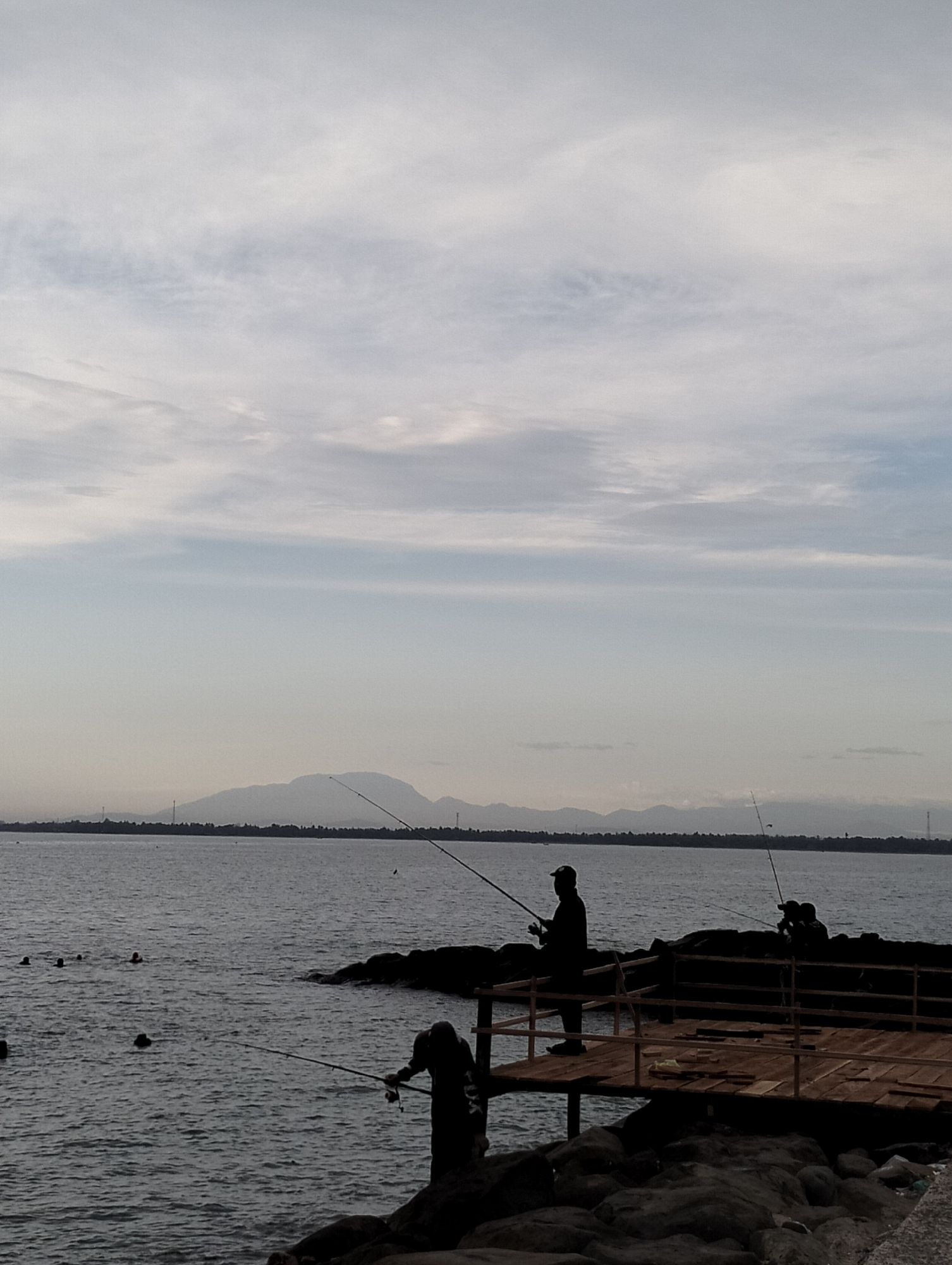 This image captures a tranquil coastal scene in Lhokseumawe, Indonesia, likely during late afternoon or early evening under an overcast or partly cloudy sky, creating a soft, diffused light.
In the foreground, the primary subjects are several people engaged in leisure activities. Three individuals are silhouetted, fishing from the shore and a wooden platform. One fisherman stands on the wooden platform, which appears to be under construction or repair, holding a fishing rod extended towards the water. Another fisherman is positioned on the dark, rugged rocks below the platform, also with a fishing rod. A third, more distant fisherman is visible on the platform to the right, with faint text that appears to read "FLUX" on their back. The platform itself is made of wooden planks and features a simple railing. To the left of the frame, several heads are visible in the water, indicating people swimming or bathing.
The midground consists of the calm, dark waters of the sea or bay. In the background, a prominent silhouette of distant mountains or large hills spans the horizon, with a narrow strip of land visible at their base. Faint details on this distant land include what might be sparse vegetation or human-made structures, and tall, slender objects resembling telecommunication towers. The upper portion of the image is dominated by a sky filled with light grey and white clouds, contributing to the overall serene and slightly hazy atmosphere. The scene evokes a sense of peaceful outdoor recreation.