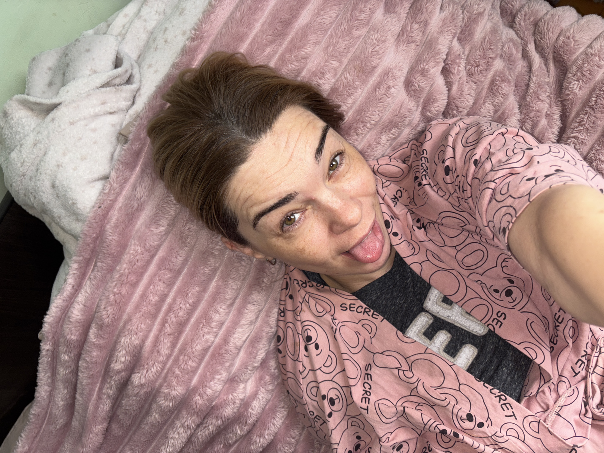A woman with brown hair and green eyes is lying on a pink blanket. She is wearing a pink robe with a teddy bear pattern and the word "SECRET" printed on it. She is sticking her tongue out at the camera. The background is a bed with a white blanket. The scene appears to be indoors, likely in a bedroom.