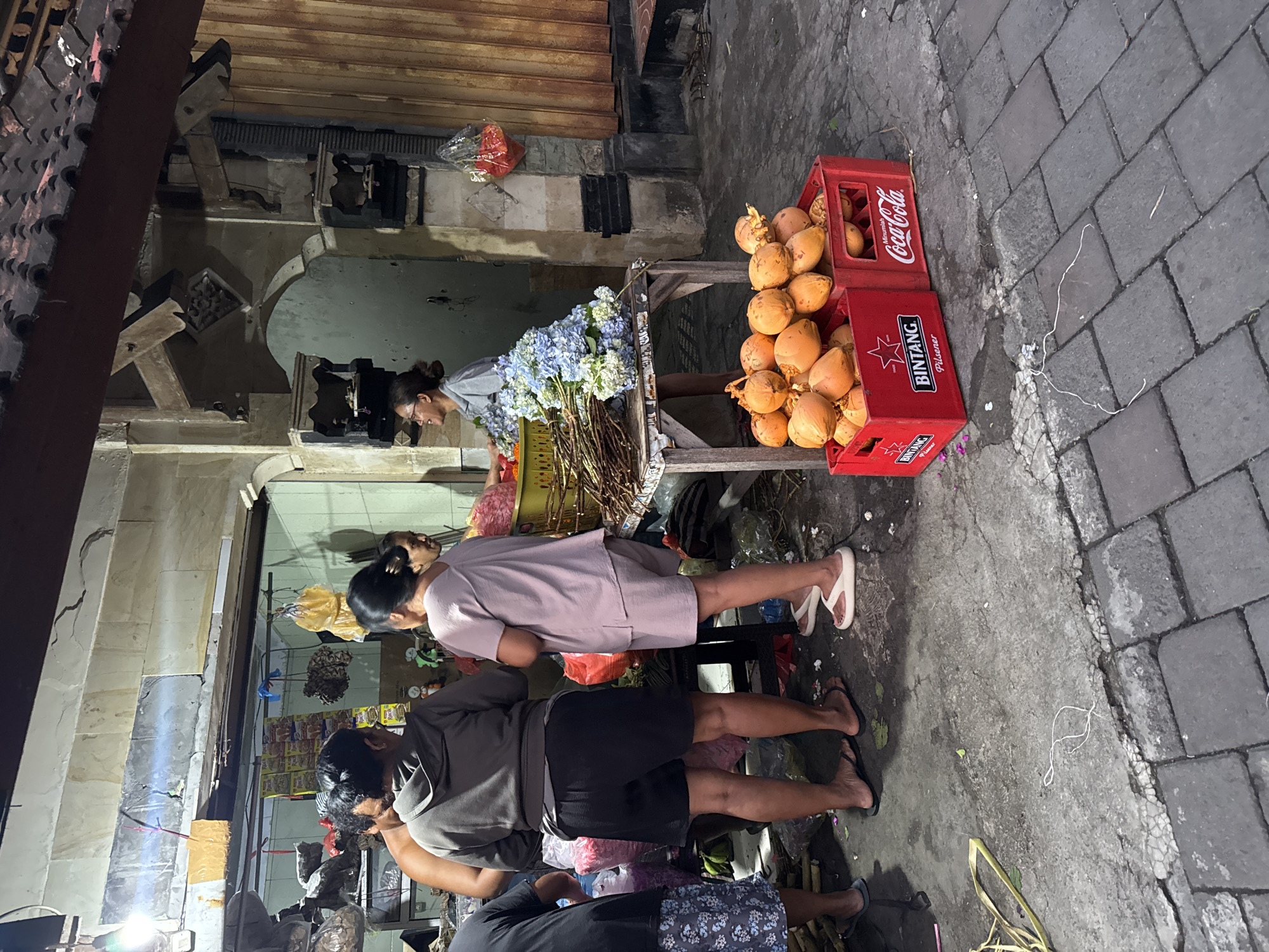 On a street in Kuta, Indonesia, a flower stand displays bouquets of blue and white flowers alongside a crate of coconuts. Four people are seen at the stand, seemingly interacting with the merchandise. The street is paved with grey bricks. The flower stand is adjacent to a building with an open doorway and what appear to be retail goods displayed inside. A Bintang beer crate and a Coca-Cola crate containing coconuts are on the street next to the stand.