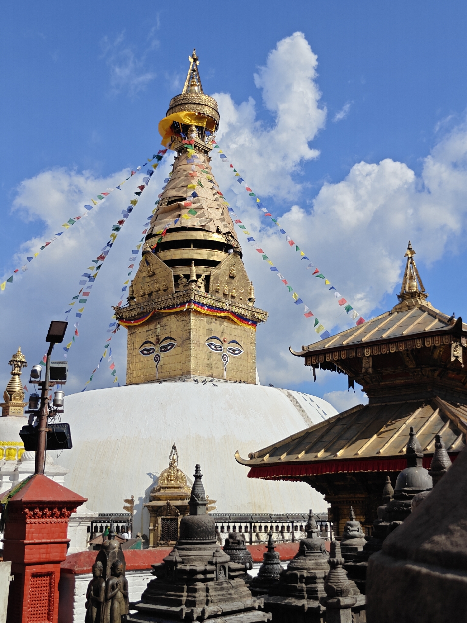 The image depicts the Swayambhunath Stupa, a UNESCO World Heritage Site in Kathmandu Metropolitan City, Nepal. The stupa, a large dome-shaped structure, is gilded with gold and features the iconic all-seeing eyes of Buddha painted on its sides. Prayer flags in various colors are strung around the stupa, fluttering in the wind against a backdrop of a blue sky with scattered white clouds. Several smaller stupas and religious structures are visible in the foreground and around the base of the main stupa. Some people are present but are too small to discern details. The scene appears to be a sunny day, with clear weather.
