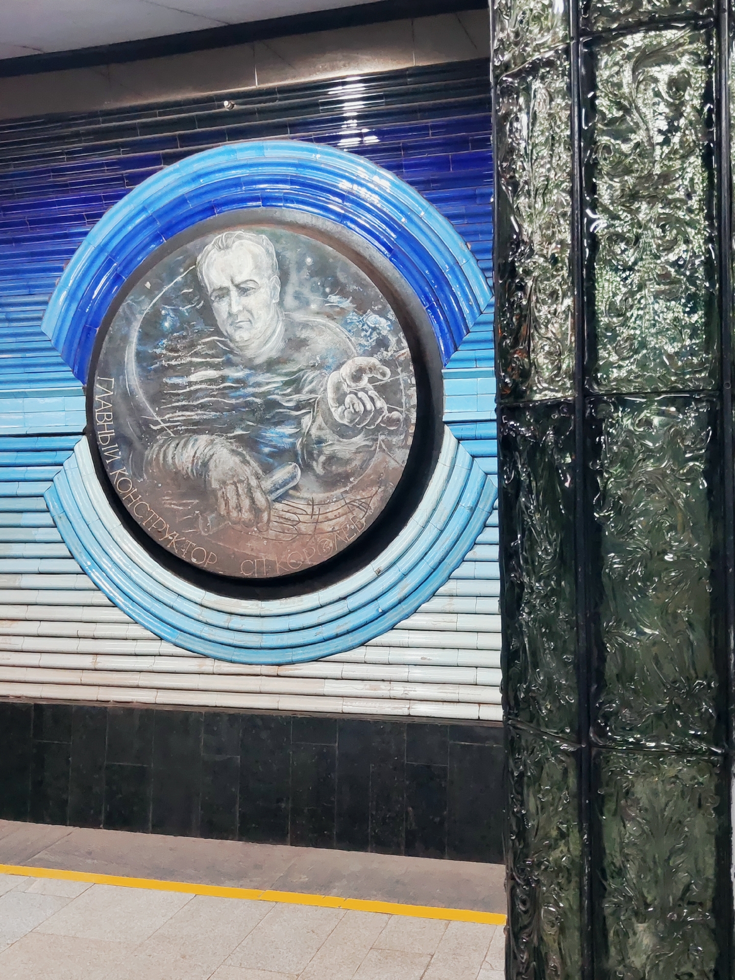 The image shows a mosaic at a metro station in Tashkent, Uzbekistan, specifically at the Kosmonavtlar (Cosmonauts) metro station. The mosaic depicts a portrait of Sergei Korolev, a prominent Soviet rocket engineer and spacecraft designer, identified by the inscription "ГЛАВНЫЙ КОНСТРУКТОР С.П. КОРОЛЕВ" (Chief Designer S.P. Korolev). He is shown in profile, looking slightly to his right, with his arms visible and holding what appears to be a schematic or blueprint. The portrait is circular and set against a background of blue and white tiles, arranged in concentric semi-circles. To the right of the mosaic, a decorative pillar made of dark green, textured glass tiles is visible, reflecting light and the surrounding environment. The station platform, with a yellow safety line, is visible in the foreground. The overall impression is of a tribute to space exploration and its pioneers.