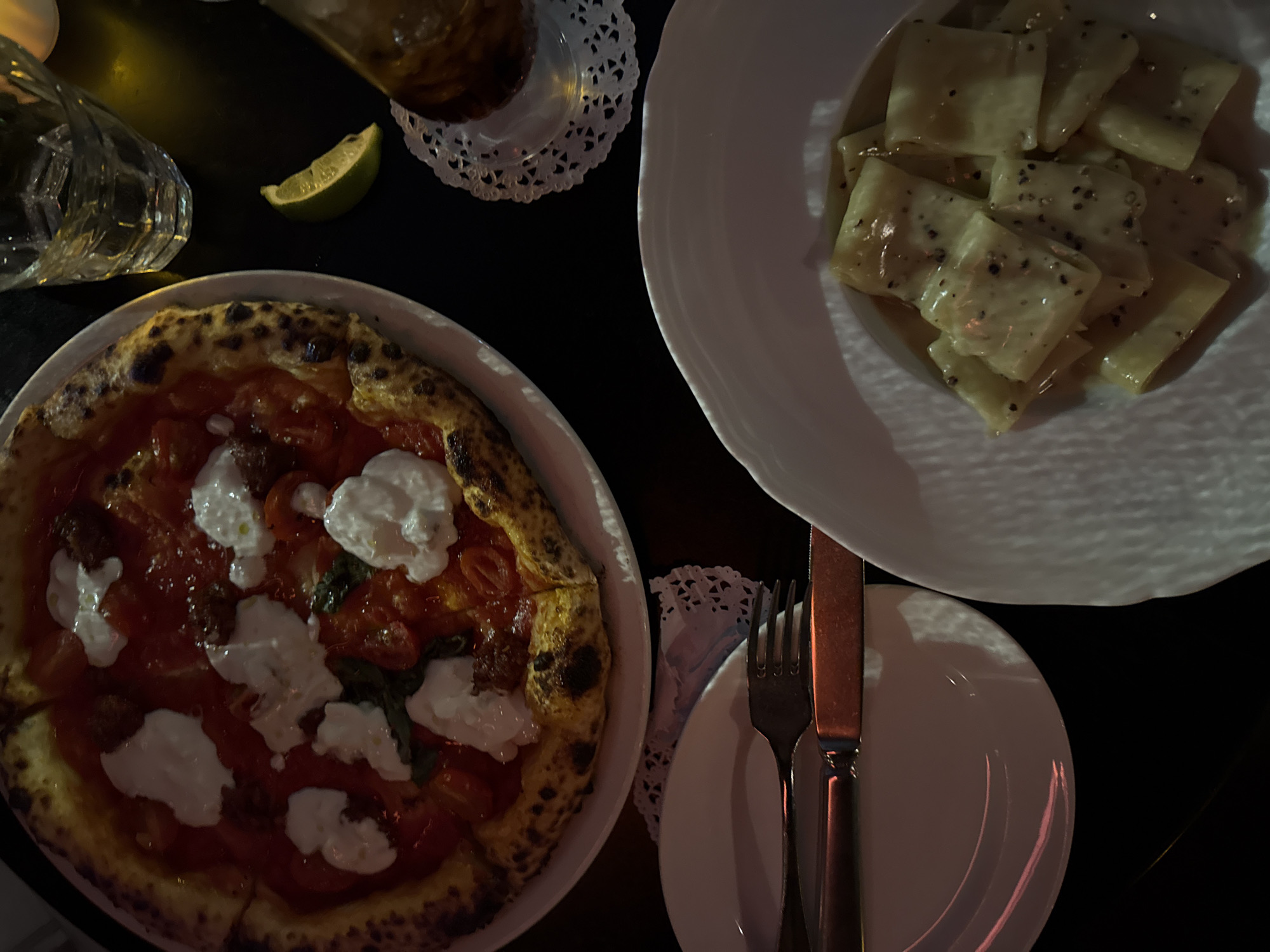 The image displays a top-down view of a dining table scene in New York, United States of America. A large Neapolitan-style pizza, characterized by a blistered crust, red tomato sauce, several dollops of white cheese (e.g., burrata or fresh mozzarella), and visible basil leaves, occupies a white circular plate in the upper left. To the right of the pizza, a clear, faceted drinking glass containing a transparent liquid and ice rests on the dark table surface, adjacent to a single green lime wedge. Further right, a second, partially visible glass holding a dark brown beverage is positioned on a white, lace-patterned paper doily. In the lower right quadrant, a white bowl with a fluted rim contains rectangular pasta shapes (e.g., paccheri) coated in a light-colored sauce with visible black pepper flakes. The lower left section features an empty white circular plate, upon which a fork and a knife, both with reddish-brown handles, are placed. This plate also rests on a white, lace-patterned paper doily. The scene is illuminated by dim, warm lighting, casting reflections on the dishware and cutlery.