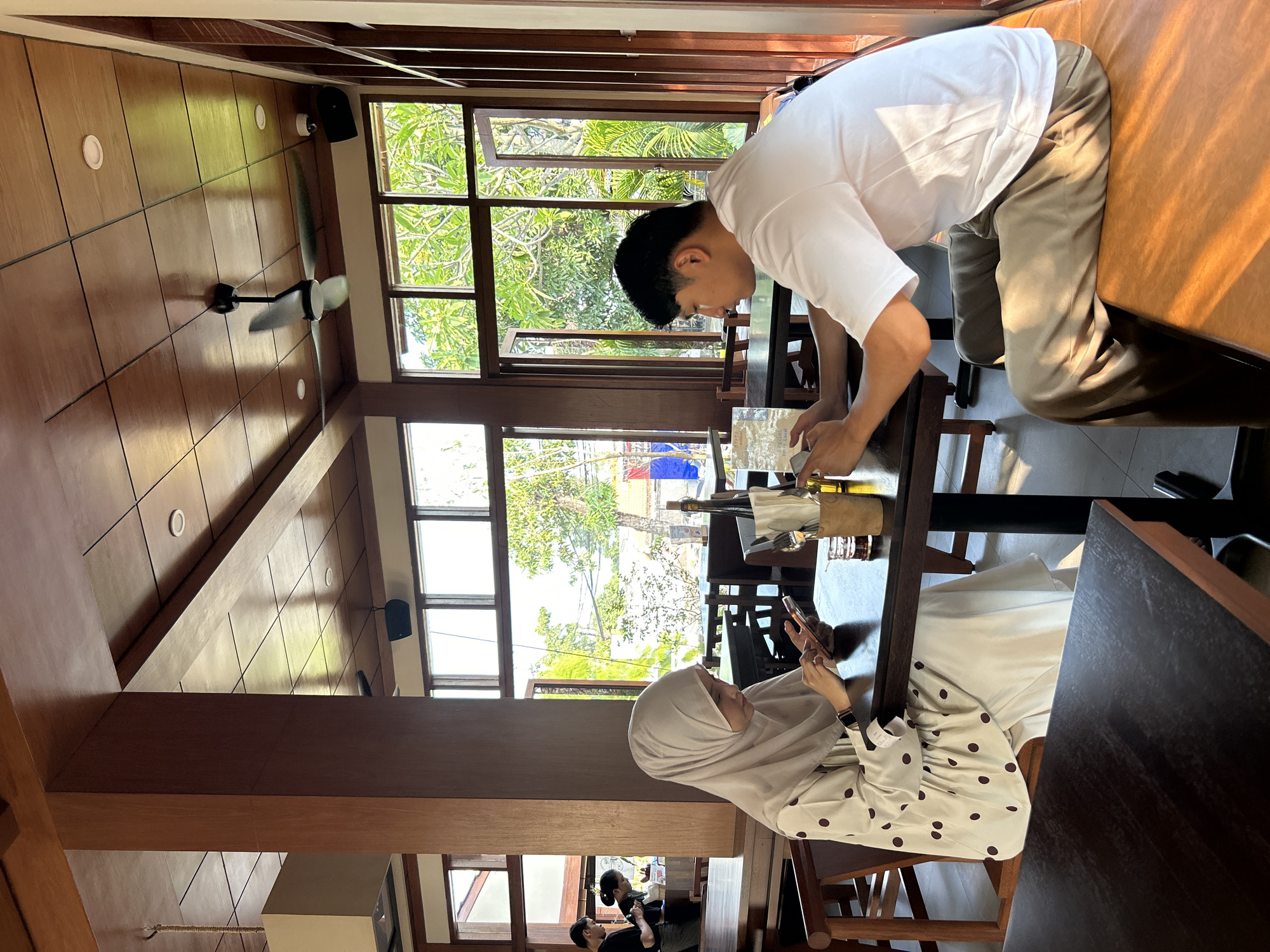 In a restaurant in North Kuta, Indonesia, a young woman wearing a beige hijab and a white polka dot dress sits at a dark wooden table, looking at her phone. Across from her, a young man in a white t-shirt and beige pants also looks at his phone. The restaurant has wooden ceilings and pillars, and large windows offer a view of lush green trees. A ceiling fan is visible on the ceiling.