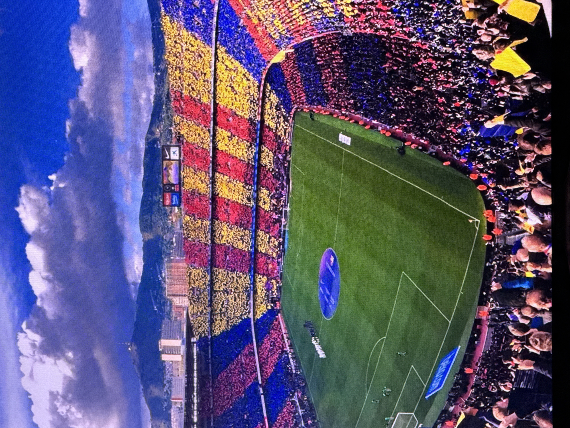 This image depicts a wide-angle view of a packed football stadium, likely during a match or pre-game ceremony. The stadium's stands are filled with spectators who are holding up colored cards to create a mosaic. The mosaic displays a pattern of blue, red, and yellow stripes, representing the colors of FC Barcelona, suggesting the stadium is Camp Nou in Barcelona, Spain, not Dubai.
The field of play is a vibrant green football pitch, with white lines marking the boundaries. In the center of the pitch, there's a large, circular blue graphic. A line of individuals, possibly players or officials, are standing on the field. In the background, beyond the stadium, a city skyline is visible with buildings and hills, under a blue sky with dramatic clouds. The overall impression is a grand sporting event with an enthusiastic crowd.